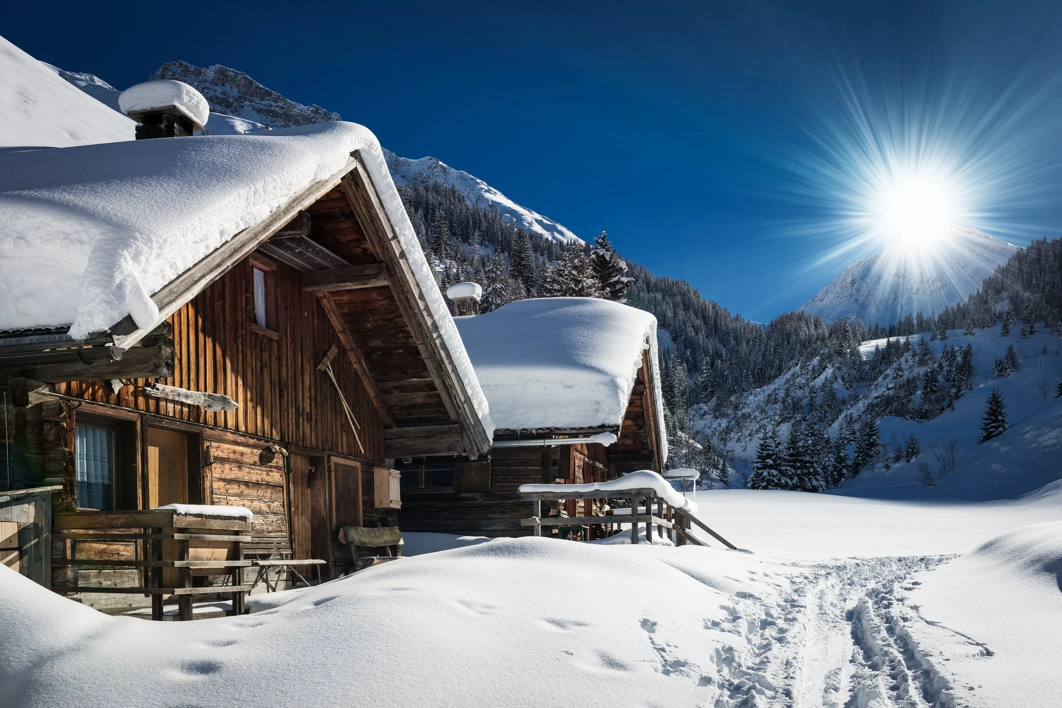 Snow-covered wooden cabin in a mountain landscape with the sun shining brightly in a clear blue sky, surrounded by pine trees and snow.