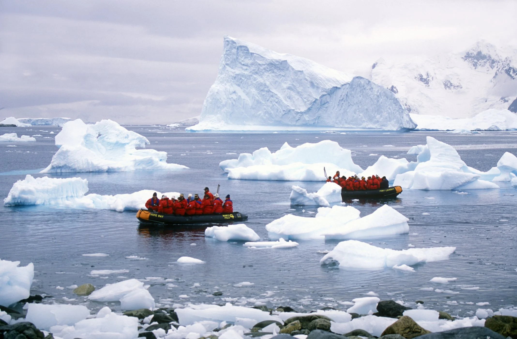 Tourists in orange life jackets riding in two black inflatable boats among icebergs in a cold, icy sea, with large ice formations and glaciers in the background.