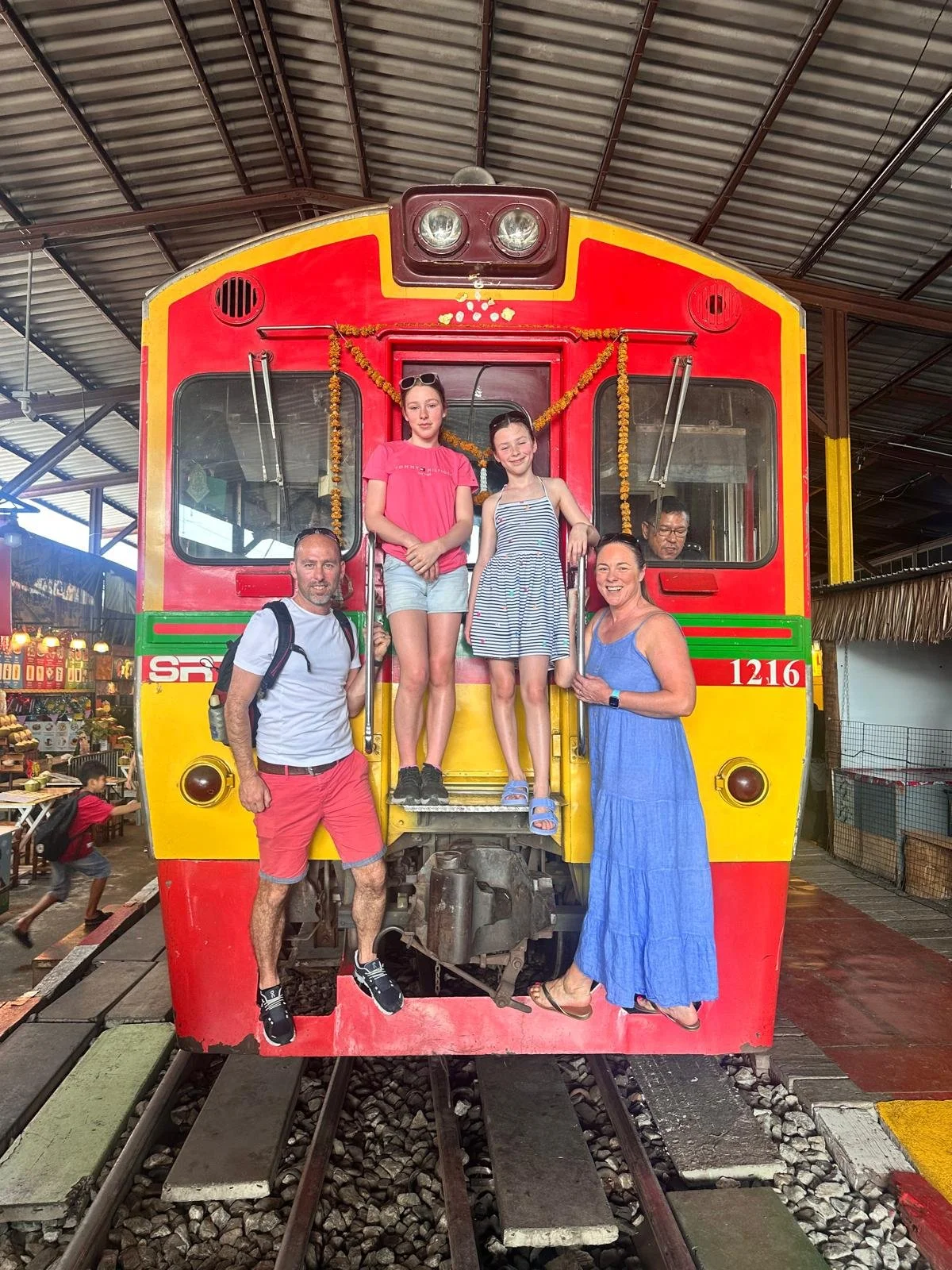 A group of five people on a colorful train at an indoor market. Two are standing on the front steps, while three are on the train's front platform, smiling for the photo.