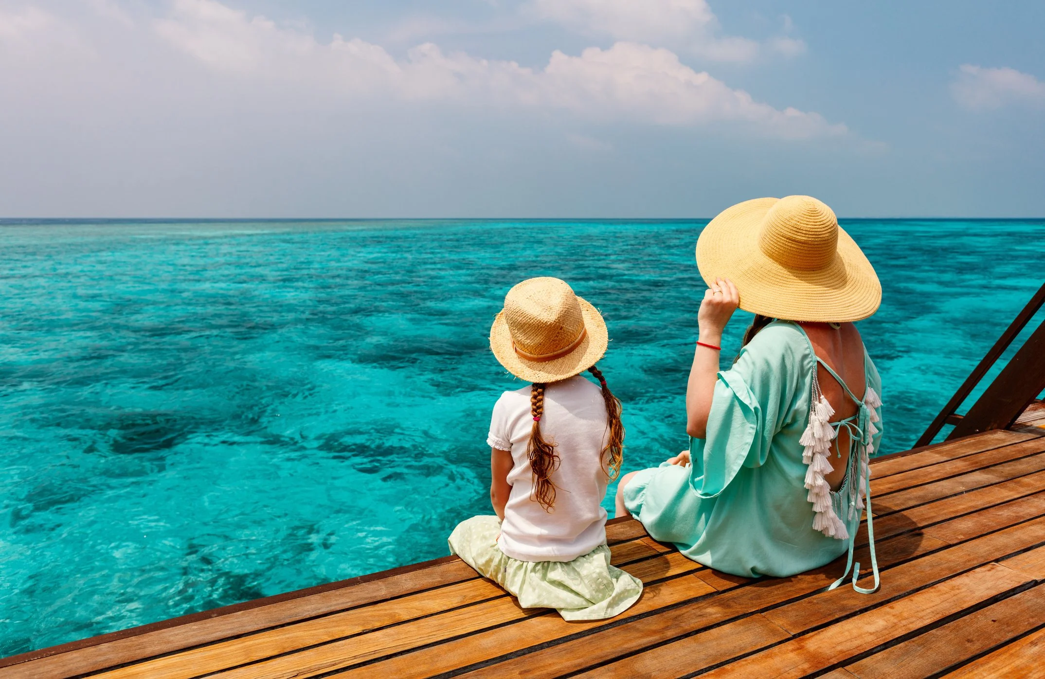 A woman and a young girl sitting on a wooden deck near the ocean, both wearing large straw sun hats and looking at the turquoise water and sky.