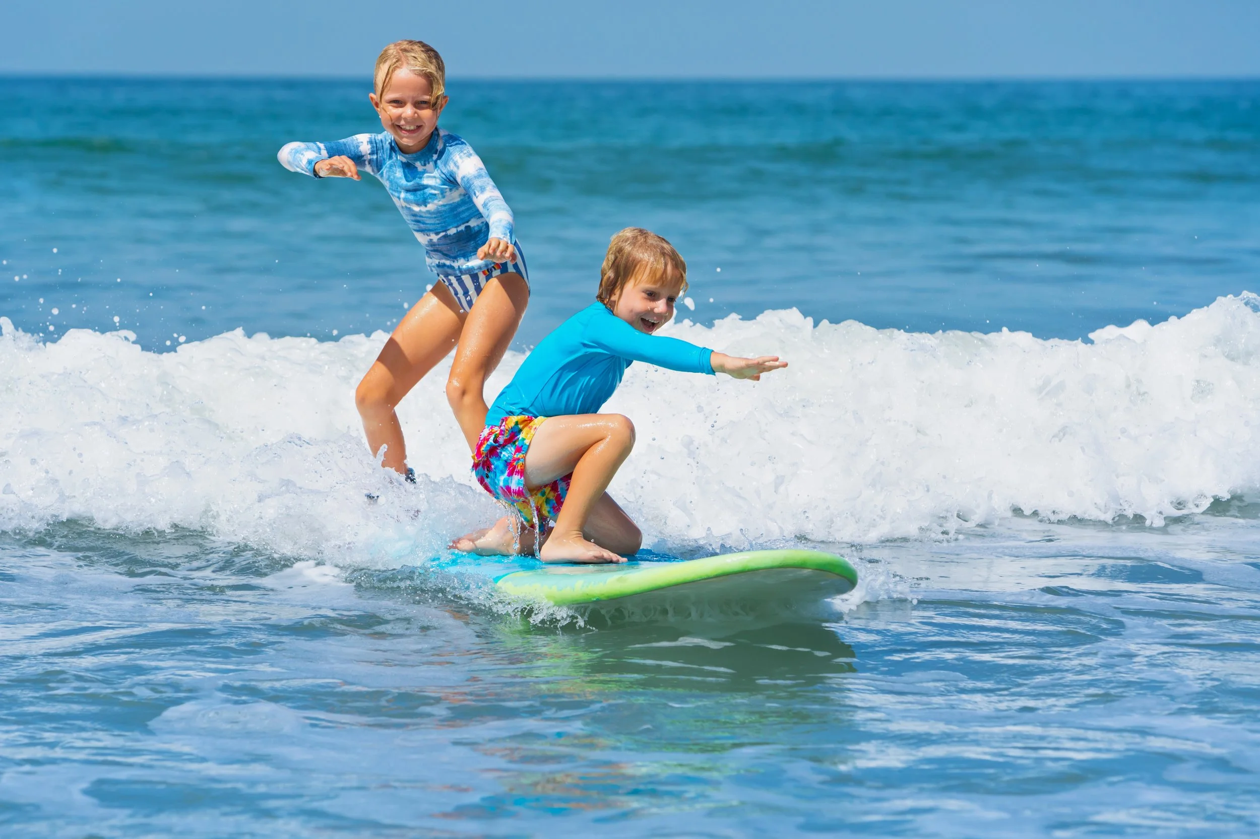 Two children, a girl and a boy, riding a surfboard on small ocean waves at the beach, smiling and enjoying the sunny day.