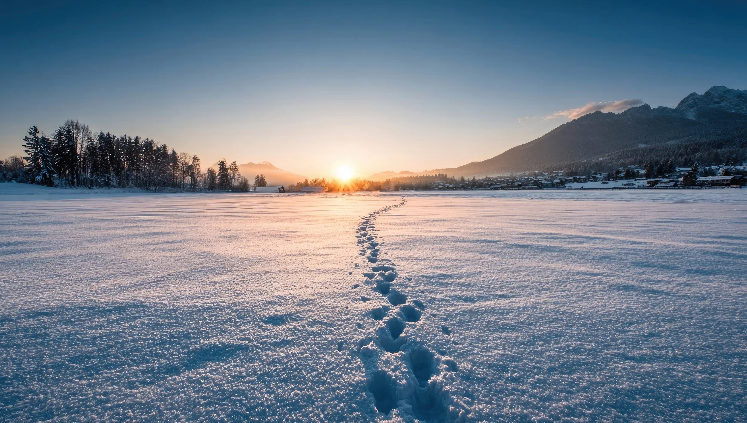 A snowy field during sunrise with footprints leading toward the horizon, mountain range in the background, and a clear sky.