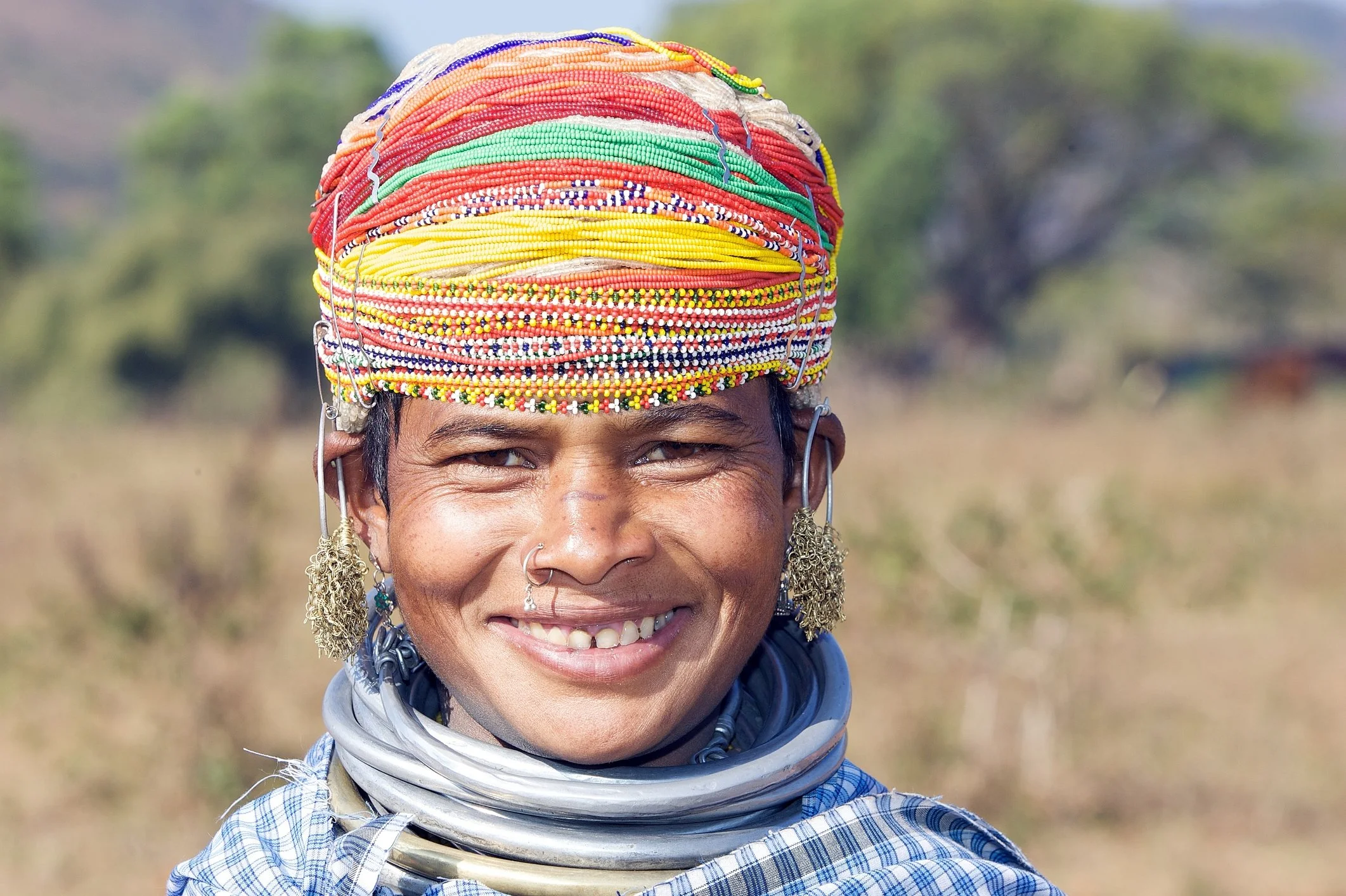 A smiling woman from the tribes of Rajasthan, India, wearing a colorful, beaded turban, large earrings, and multiple silver necklaces, with a blurred natural landscape in the background.