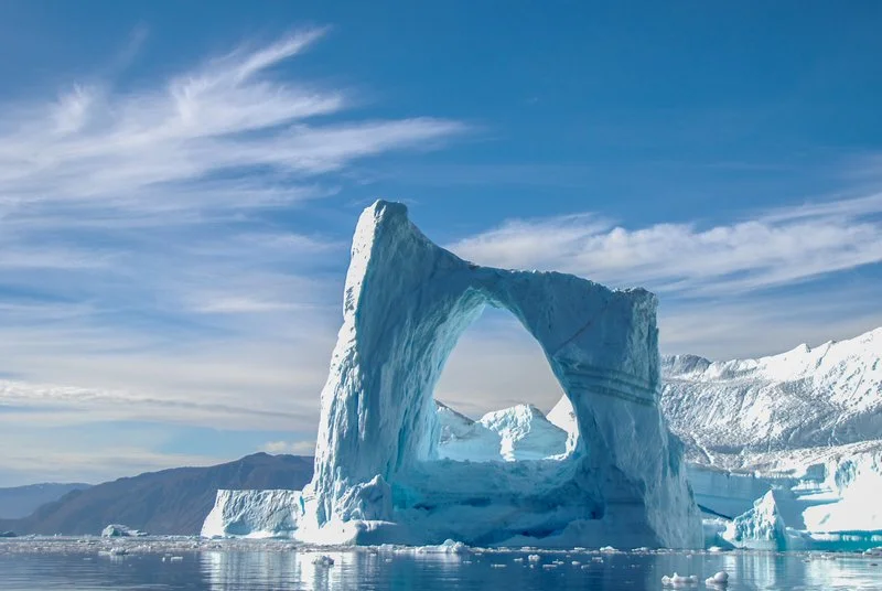 Large iceberg with a natural arch in a cold, icy ocean under a partly cloudy sky.