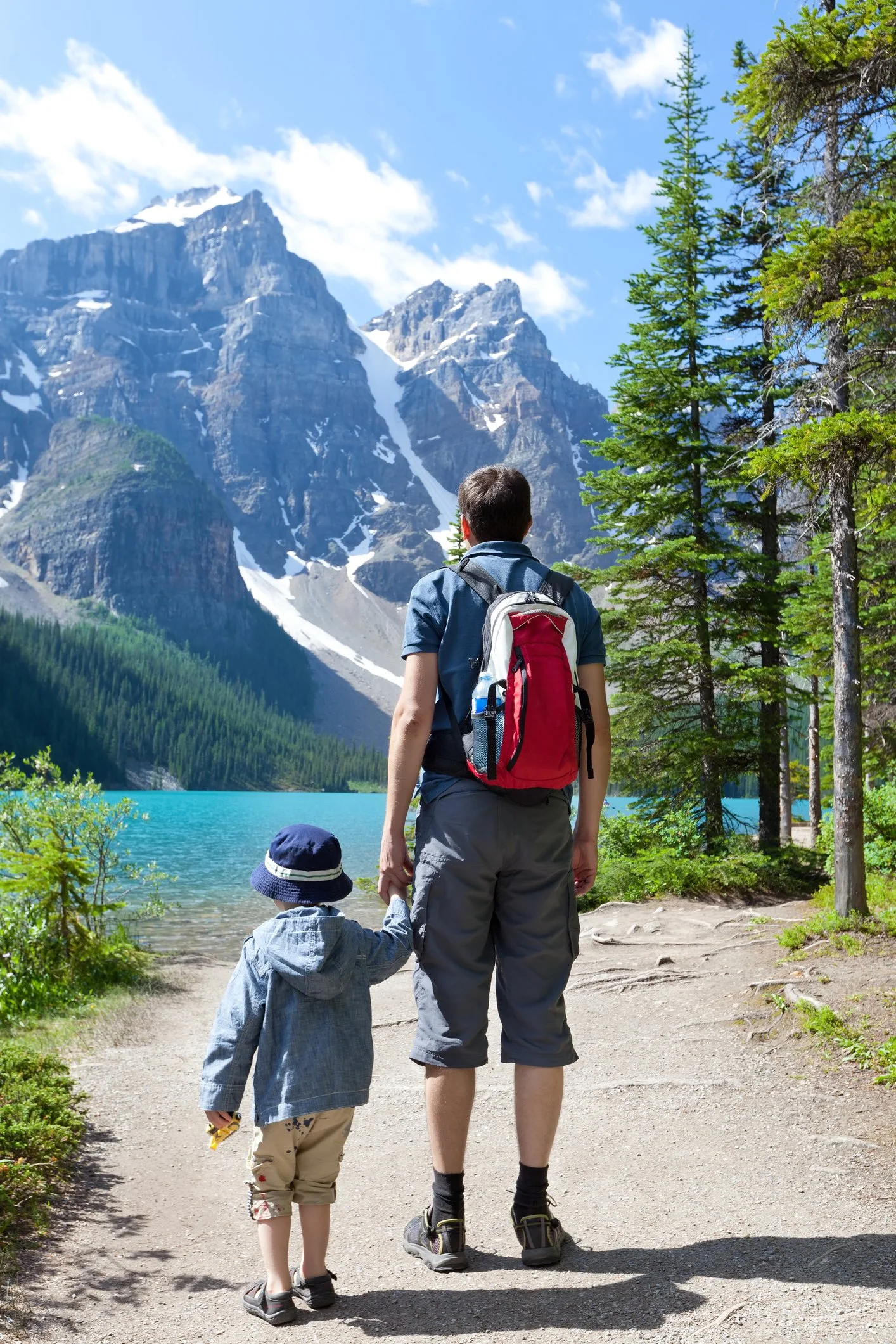 A man and a young child walking on a dirt trail beside a lake with mountains in the background. The man is wearing a backpack and holding the child's hand. The scene is outdoors during daytime with clear skies.