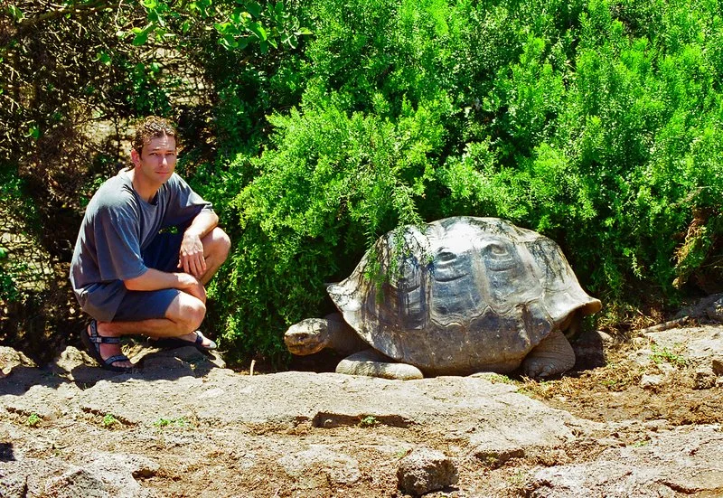 A man squatting next to a large tortoise on a dirt path with green bushes in the background.