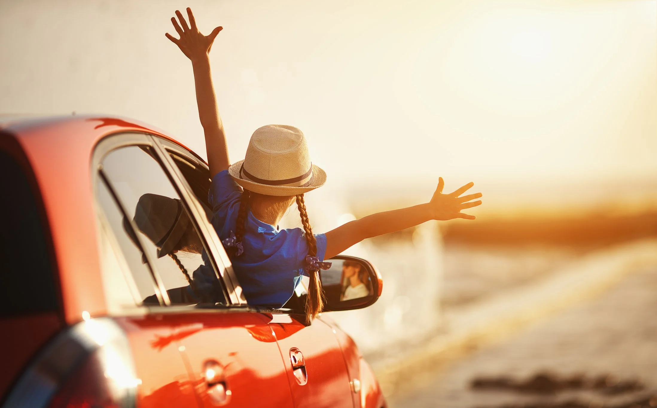 A young girl with braided hair wearing a straw hat and blue shirt leaning out of a red car window with arms outstretched during sunset at the beach.