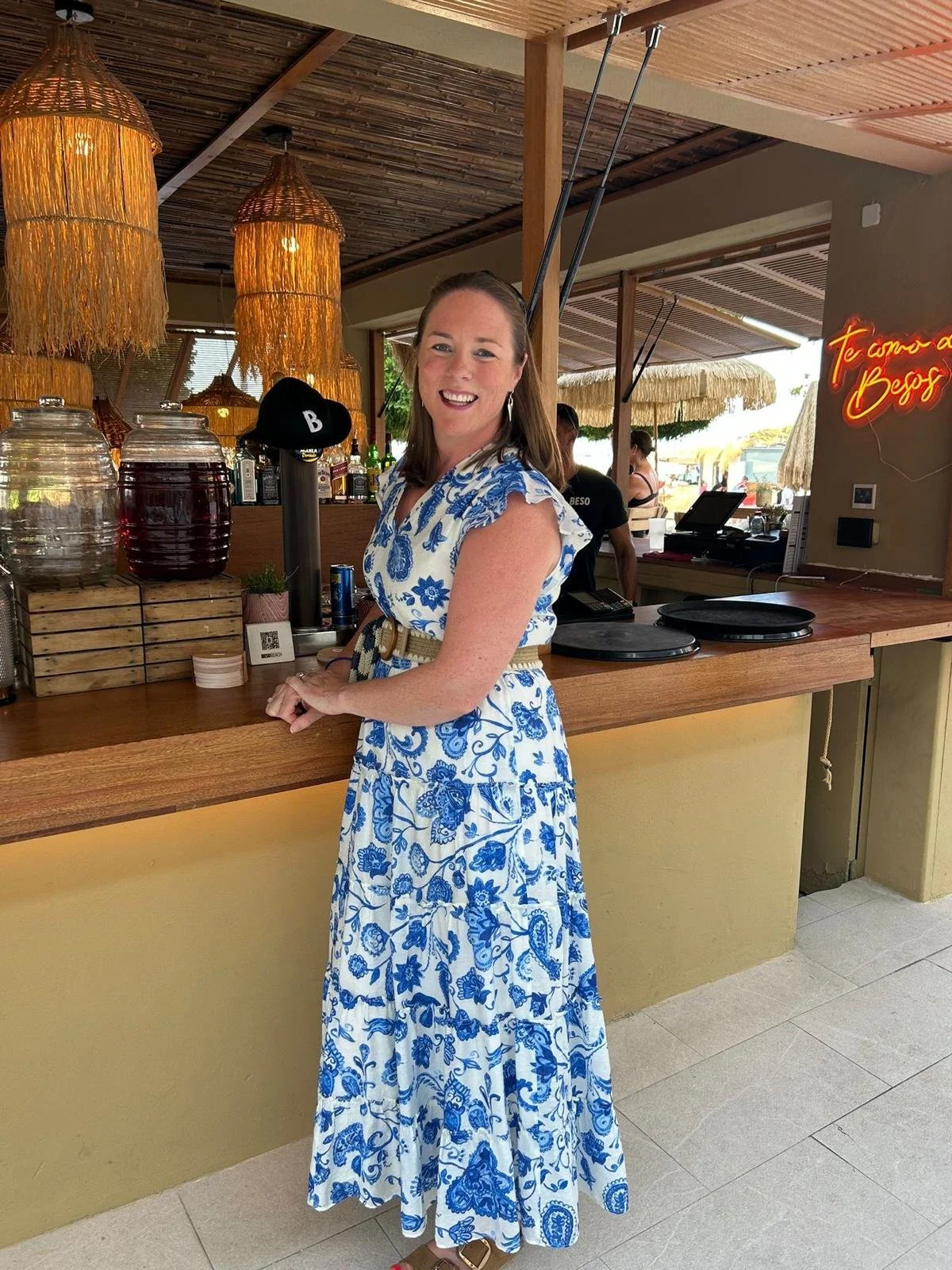 A woman in a white and blue floral dress smiling at a bar with hanging wicker lamps and large jars of drinks in a tropical open-air setting.