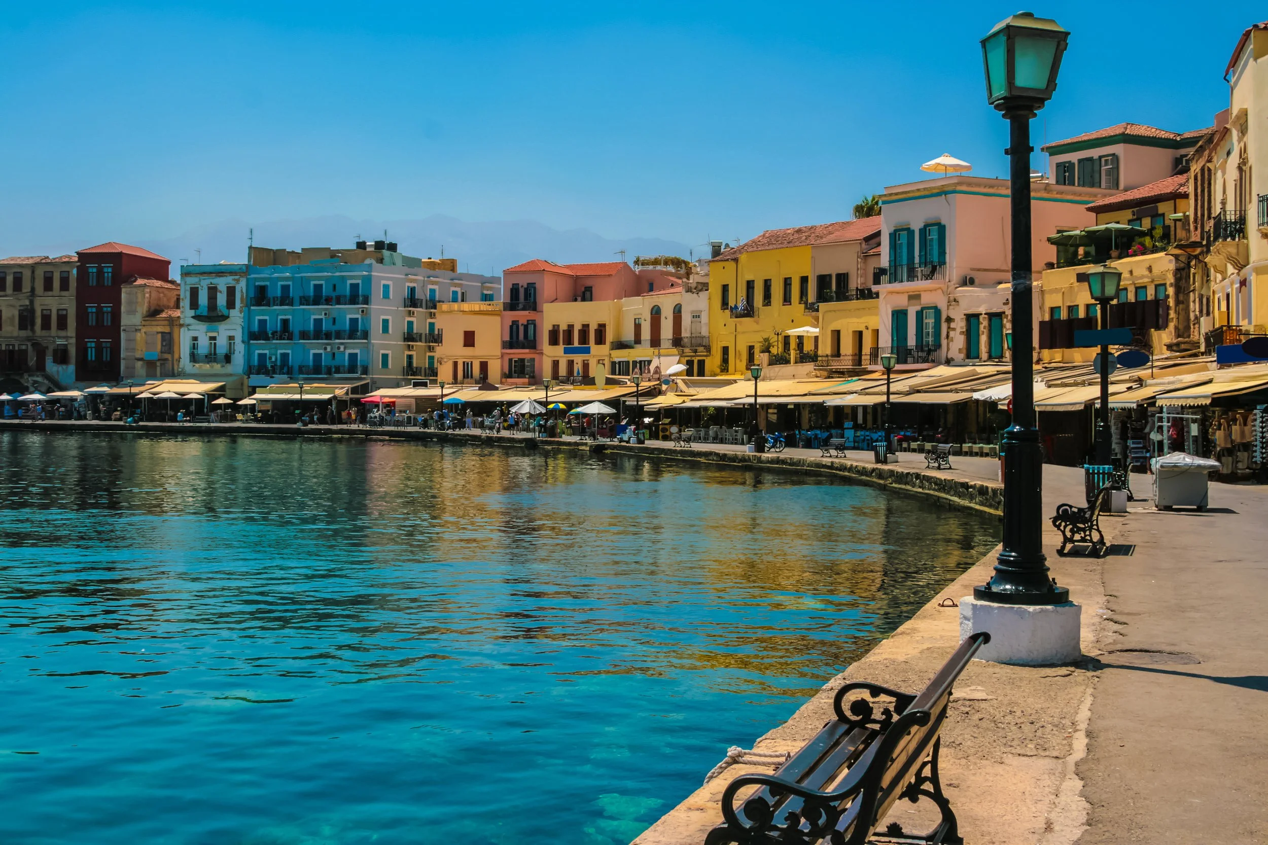 Colorful buildings along a waterfront promenade with benches, street lamps, and outdoor cafes, on a sunny day with blue sky.