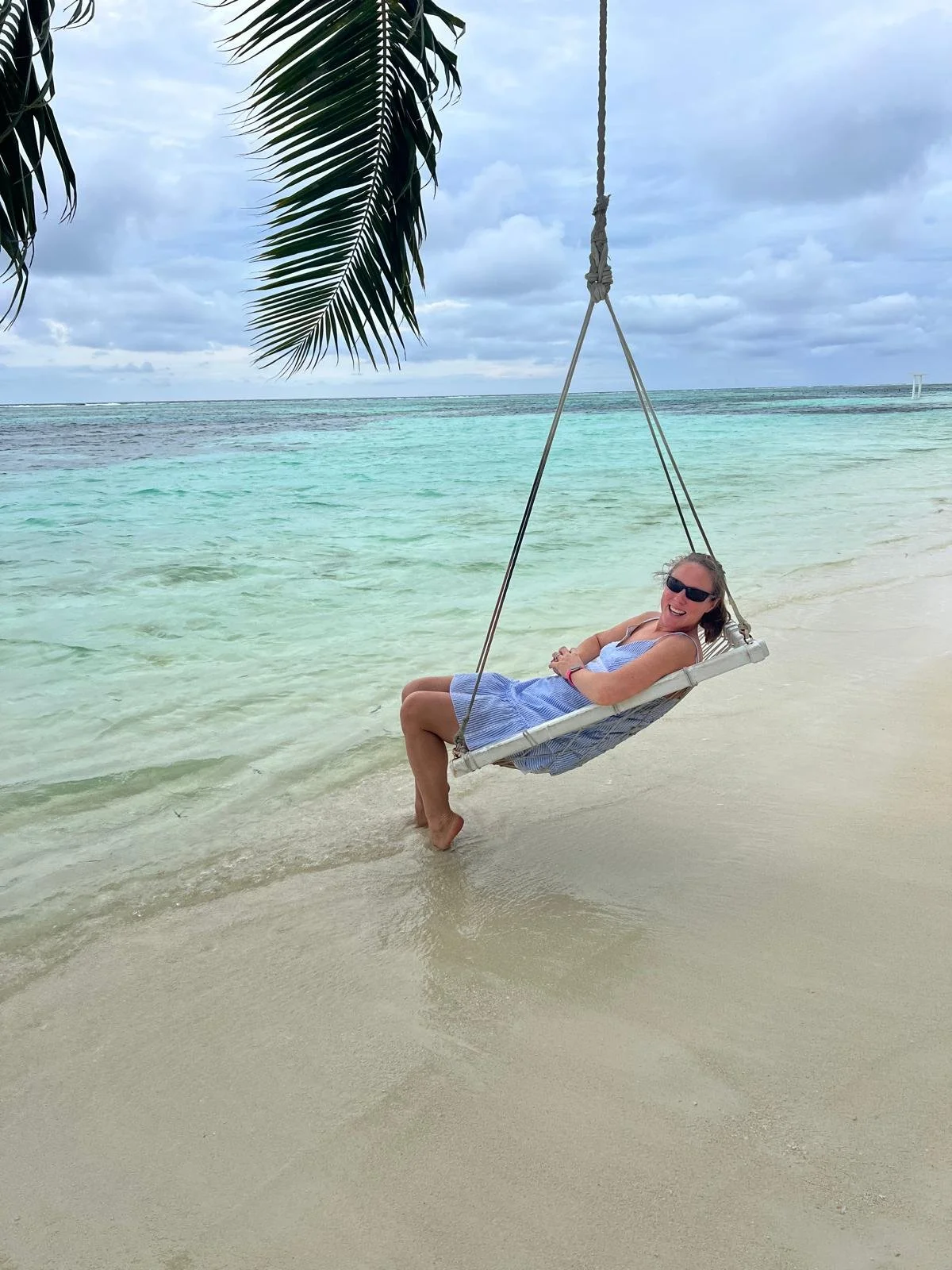 A woman in sunglasses in a blue dress lounging on a white swing hanging from a palm tree on a sandy beach, with turquoise water and cloudy sky in the background.