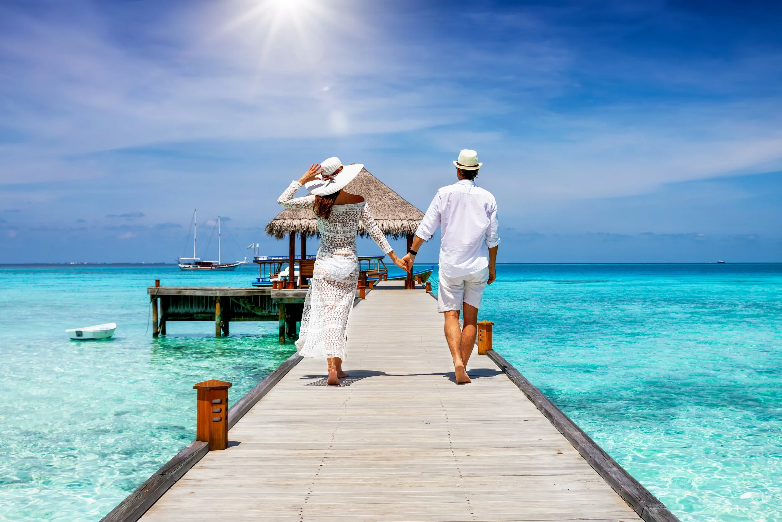 A couple holding hands walking on a wooden pier over clear turquoise water towards a thatched-roof hut, with sailboats in the background under a blue sky.