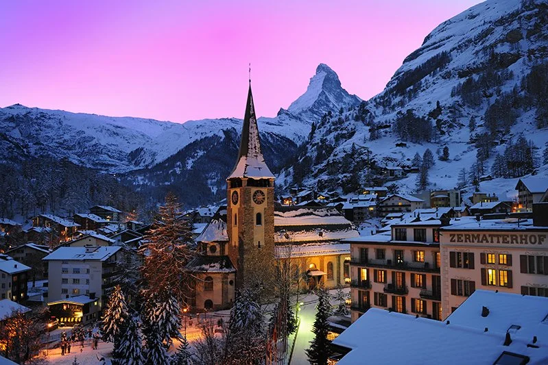 Snow-covered buildings and trees in a mountain village at dusk, with a church featuring a tall steeple and clock, and a pink and purple sky in the background.