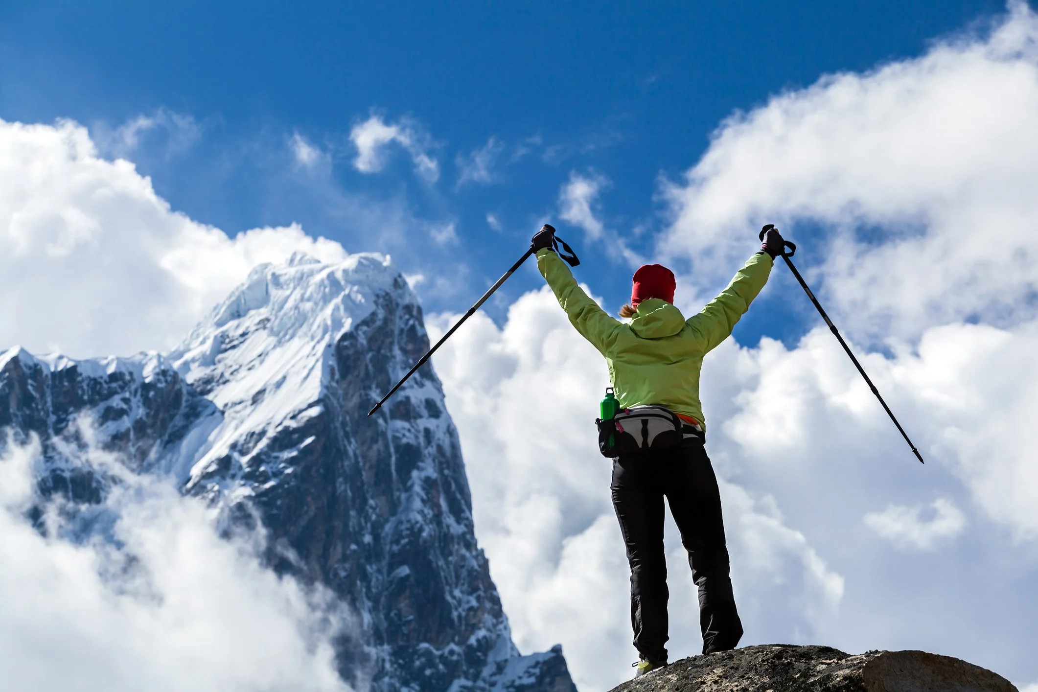 Person standing on a mountain peak with arms raised holding trekking poles, facing a snowy mountain under a partly cloudy blue sky.