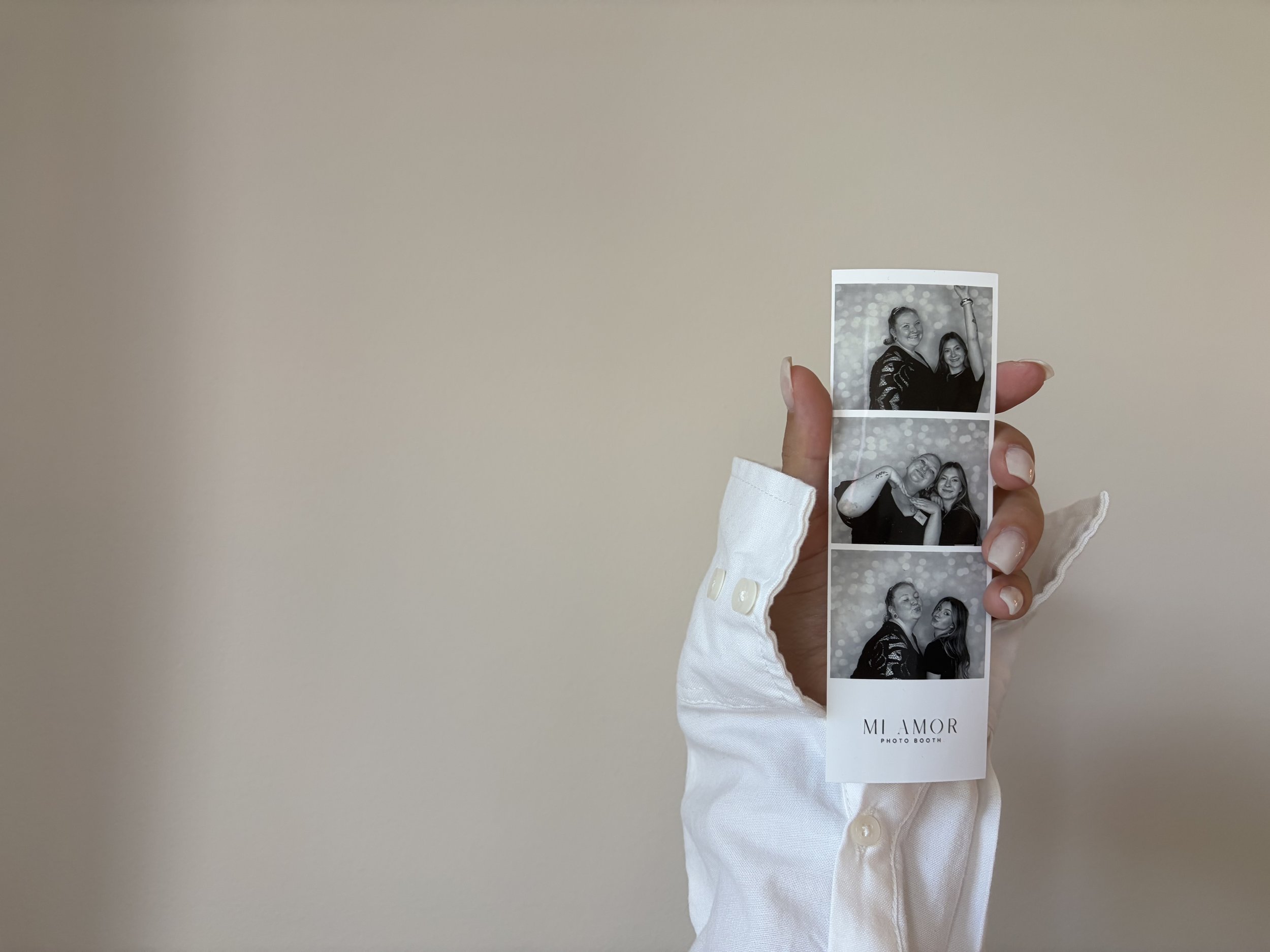 Person holding a strip of black and white photo booth pictures against a plain beige wall. The visible images show two women, one smiling and the other making a playful gesture, and both posing together.