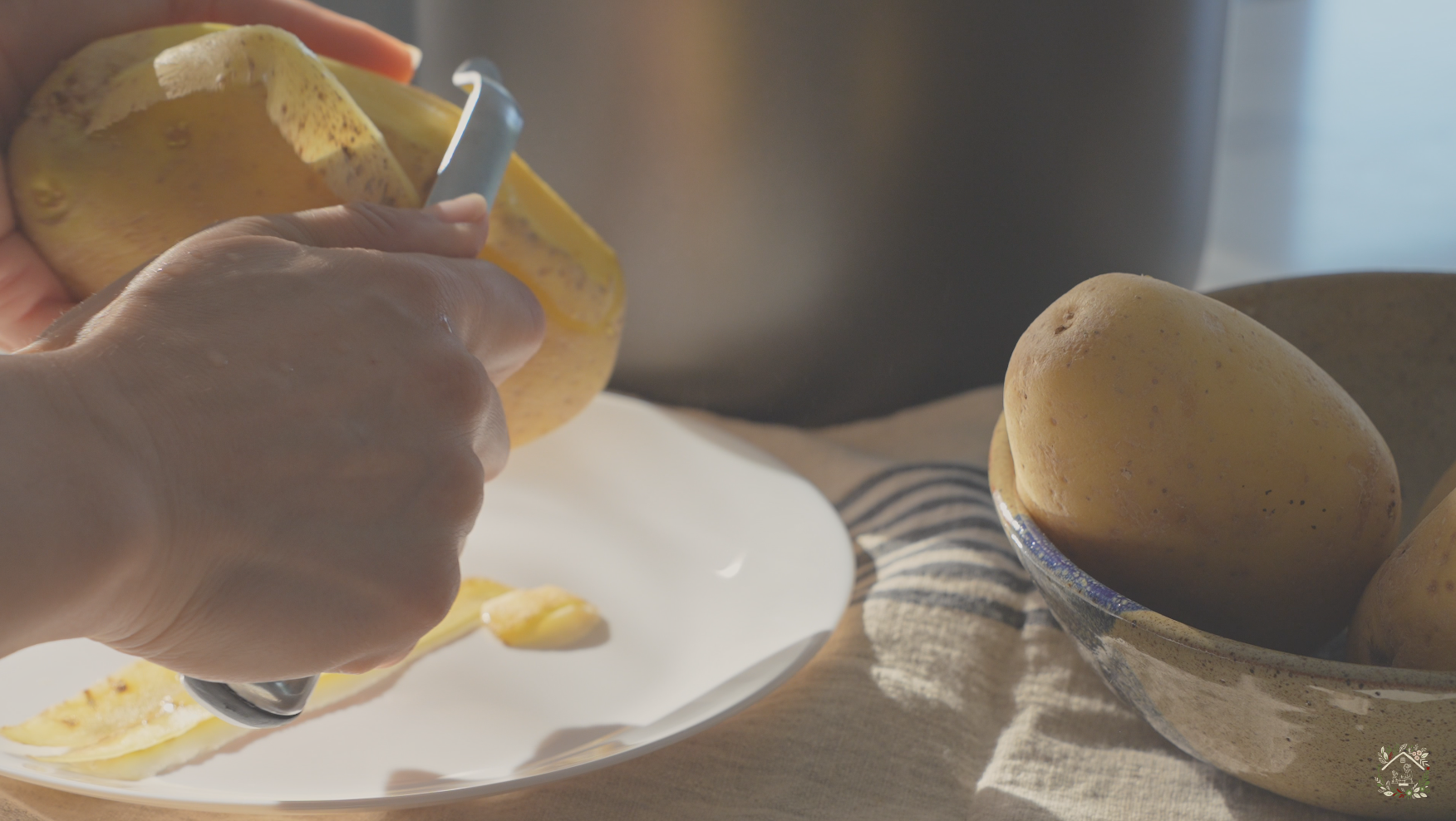 A person peeling a potato with a vegetable peeler near a white plate and a bowl of unpeeled potatoes on a cloth-covered surface.