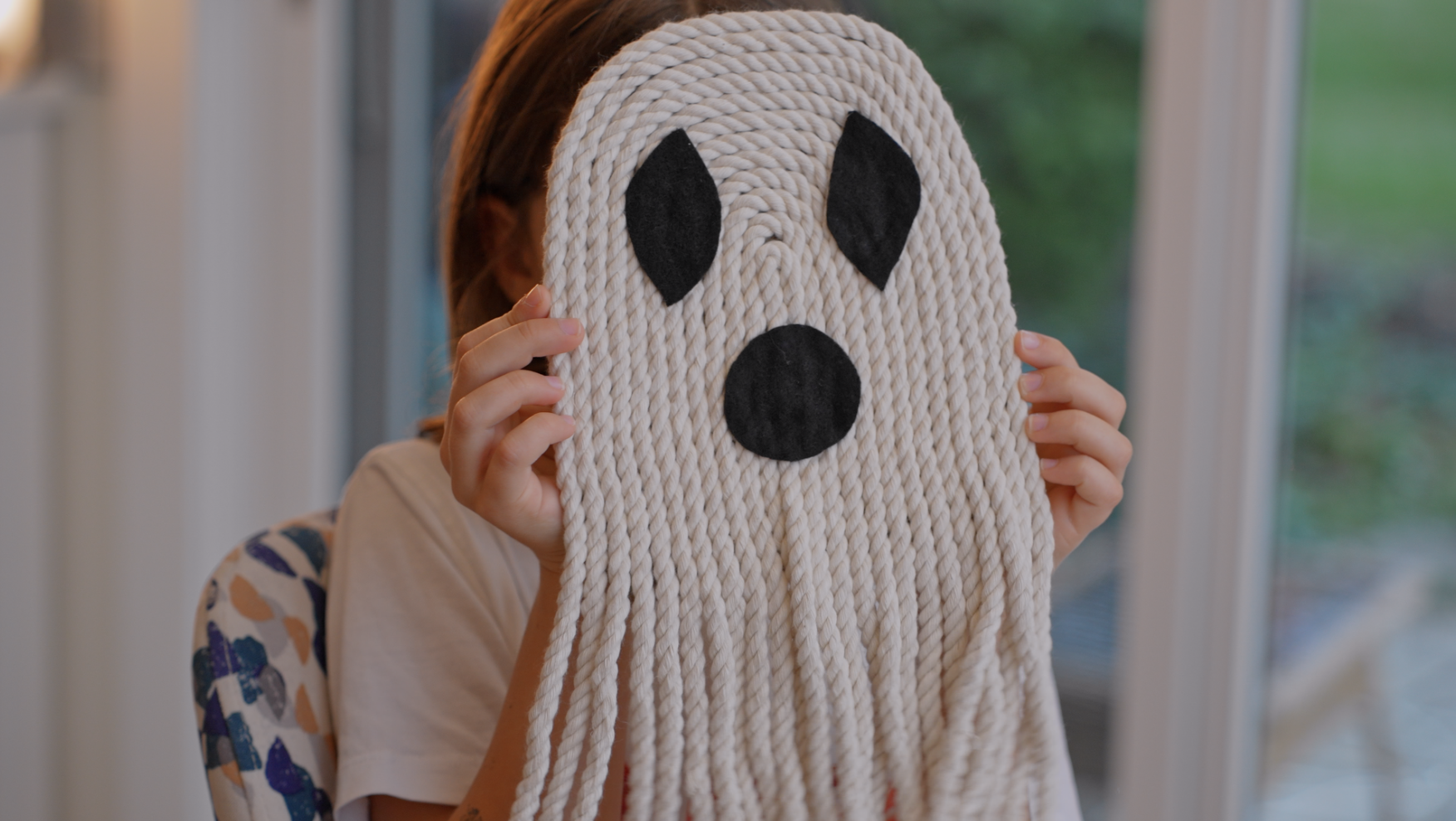 Child holding a ghost-shaped craft mask with black eyes and mouth, made of twisted yarn, in front of their face.