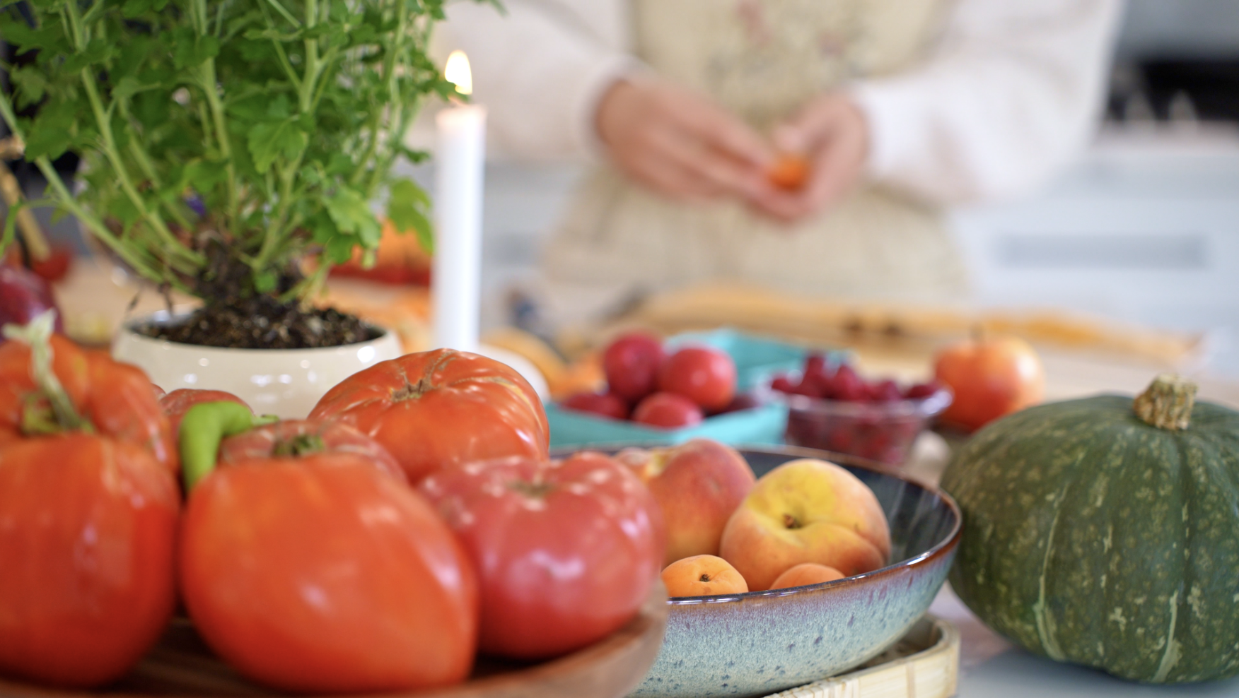 A table with various fresh vegetables and fruits, including tomatoes, peaches, a green pumpkin, and a potted plant, with a person in the background preparing food.