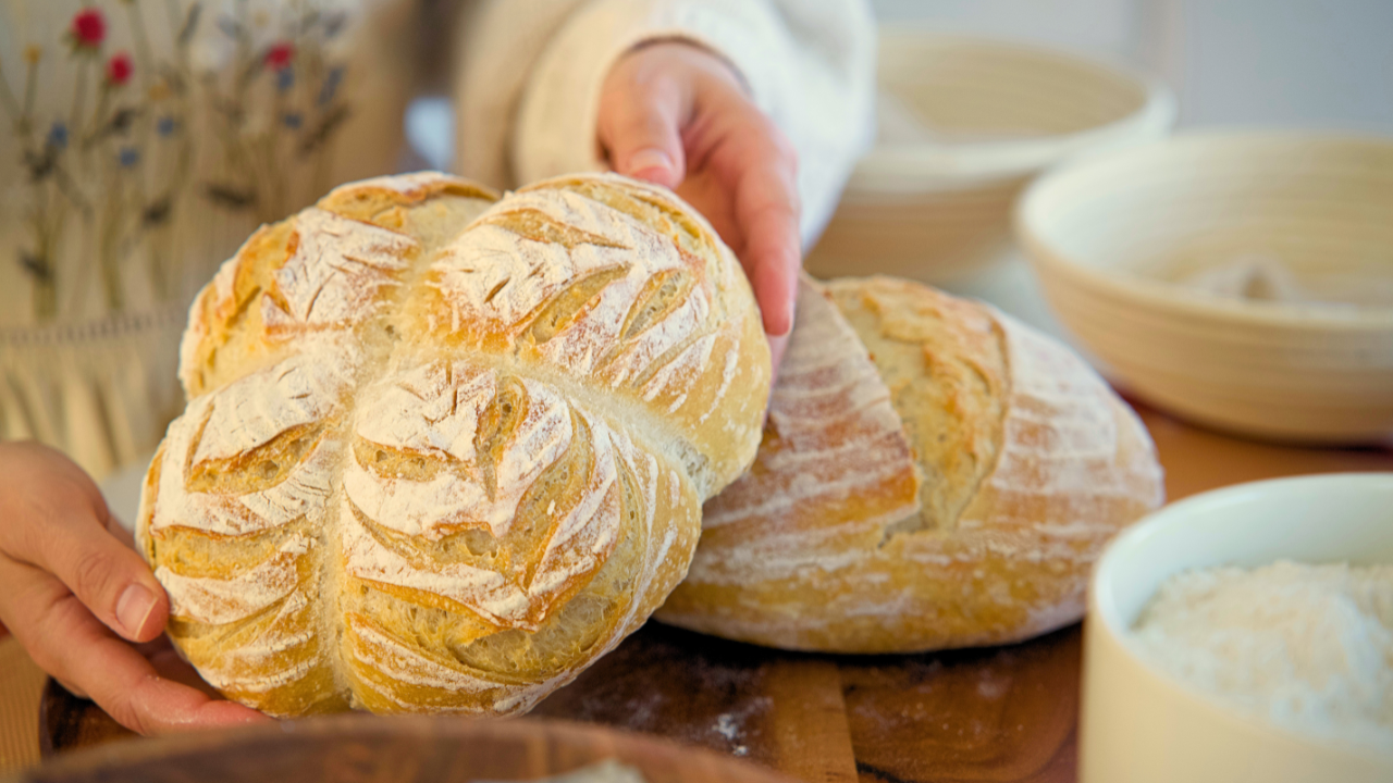 Person holding freshly baked artisanal bread in a kitchen with bowls of flour and dough in the background.