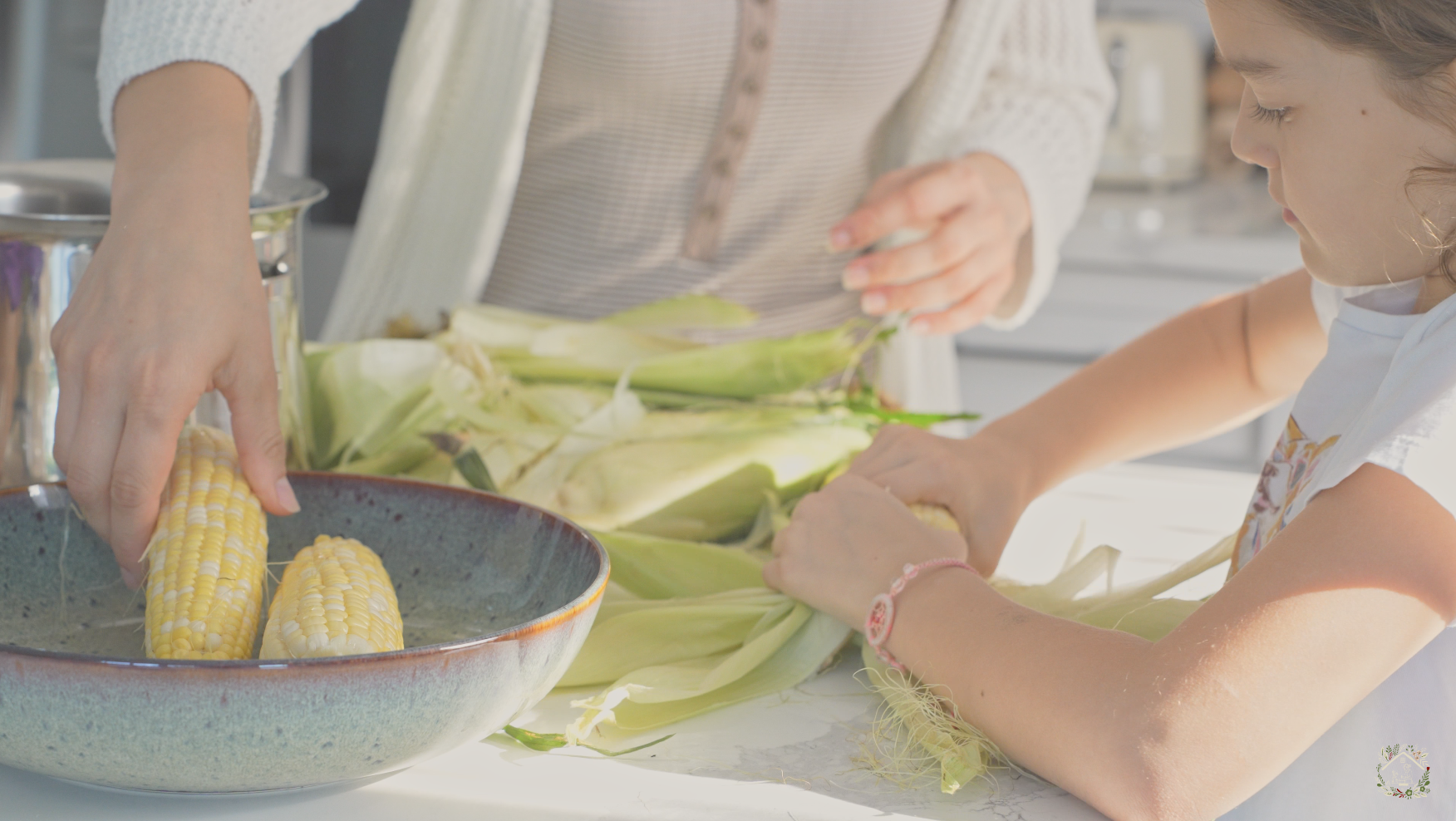 A person and a young girl shucking fresh corn on a kitchen counter, with corn husks and a bowl of corn nearby, in a well-lit kitchen.