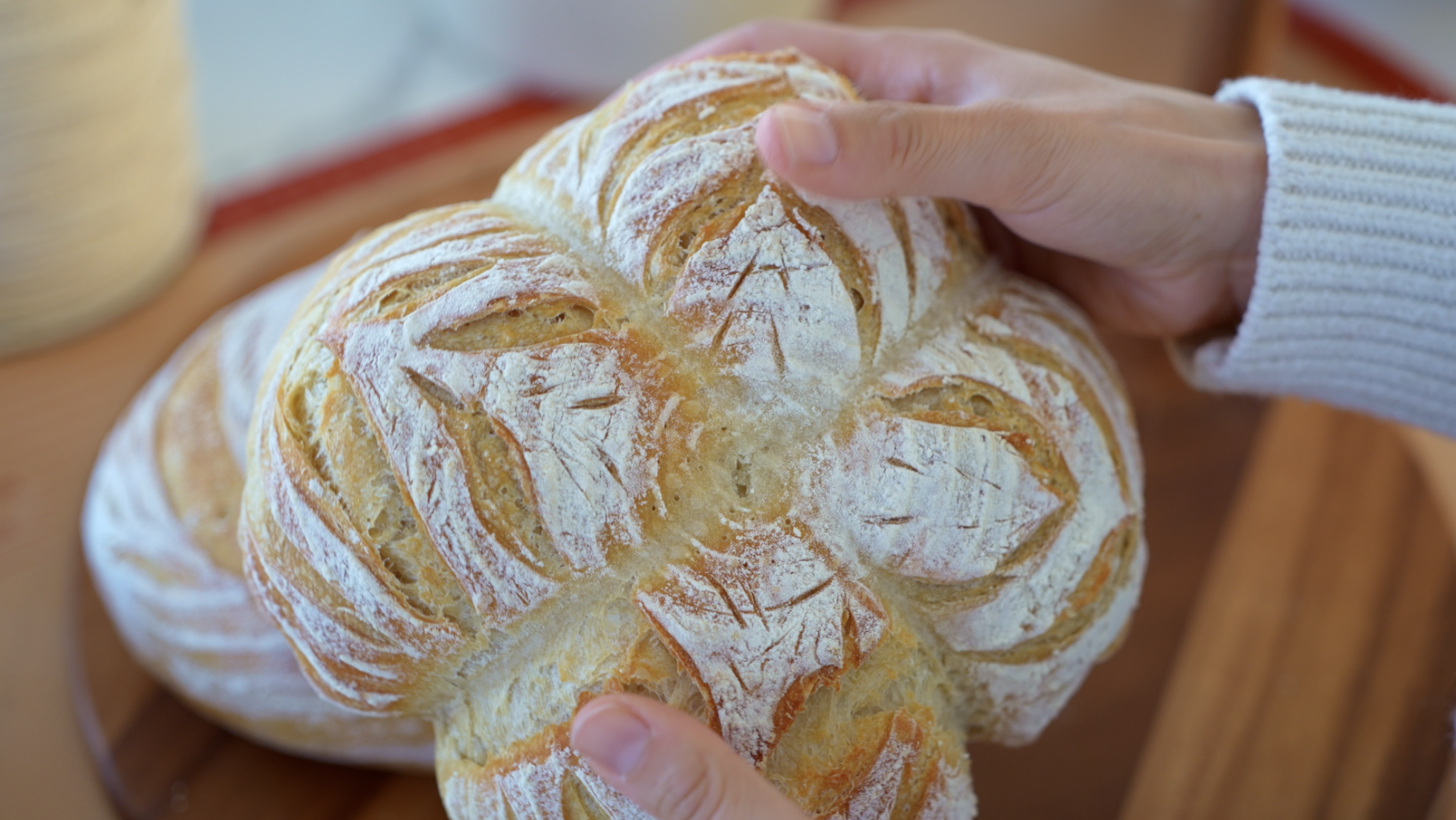 Close-up of a person's hand touching a rustic, round loaf of bread with a flour-dusted crust and intricate scoring patterns, on a wooden surface.