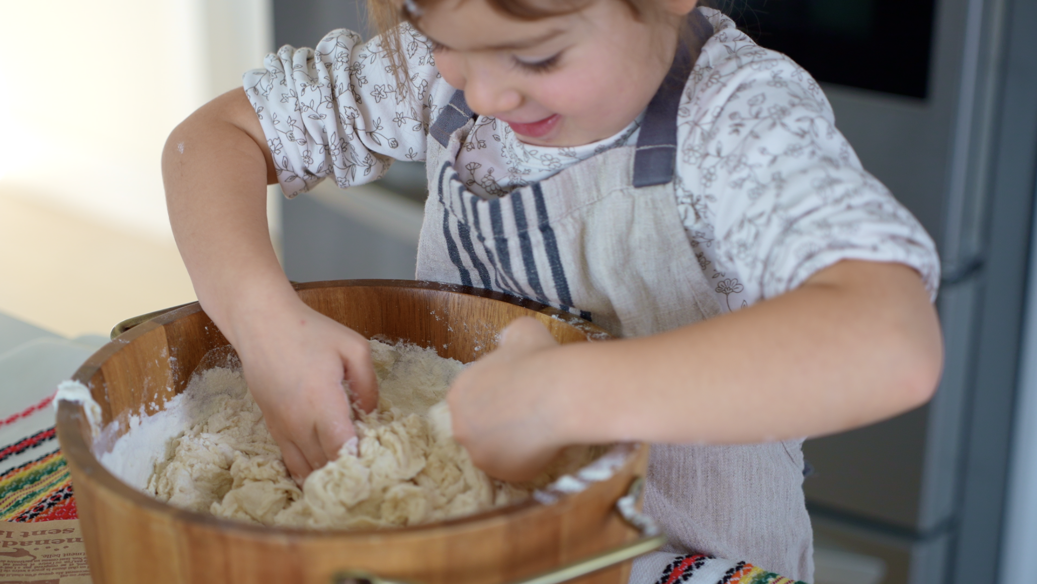 A young child in a striped apron mixing dough with hands in a wooden bowl.