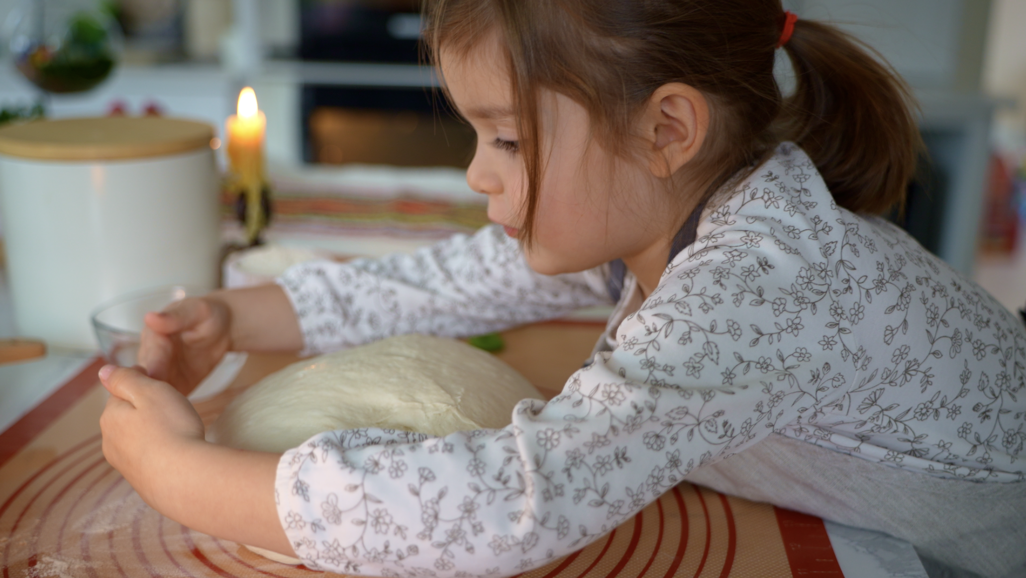 A young girl with brown hair tied in a red ponytail, wearing a white shirt with black floral pattern, is pressing her hands on a ball of dough on a kitchen counter, with a small glass bowl nearby.