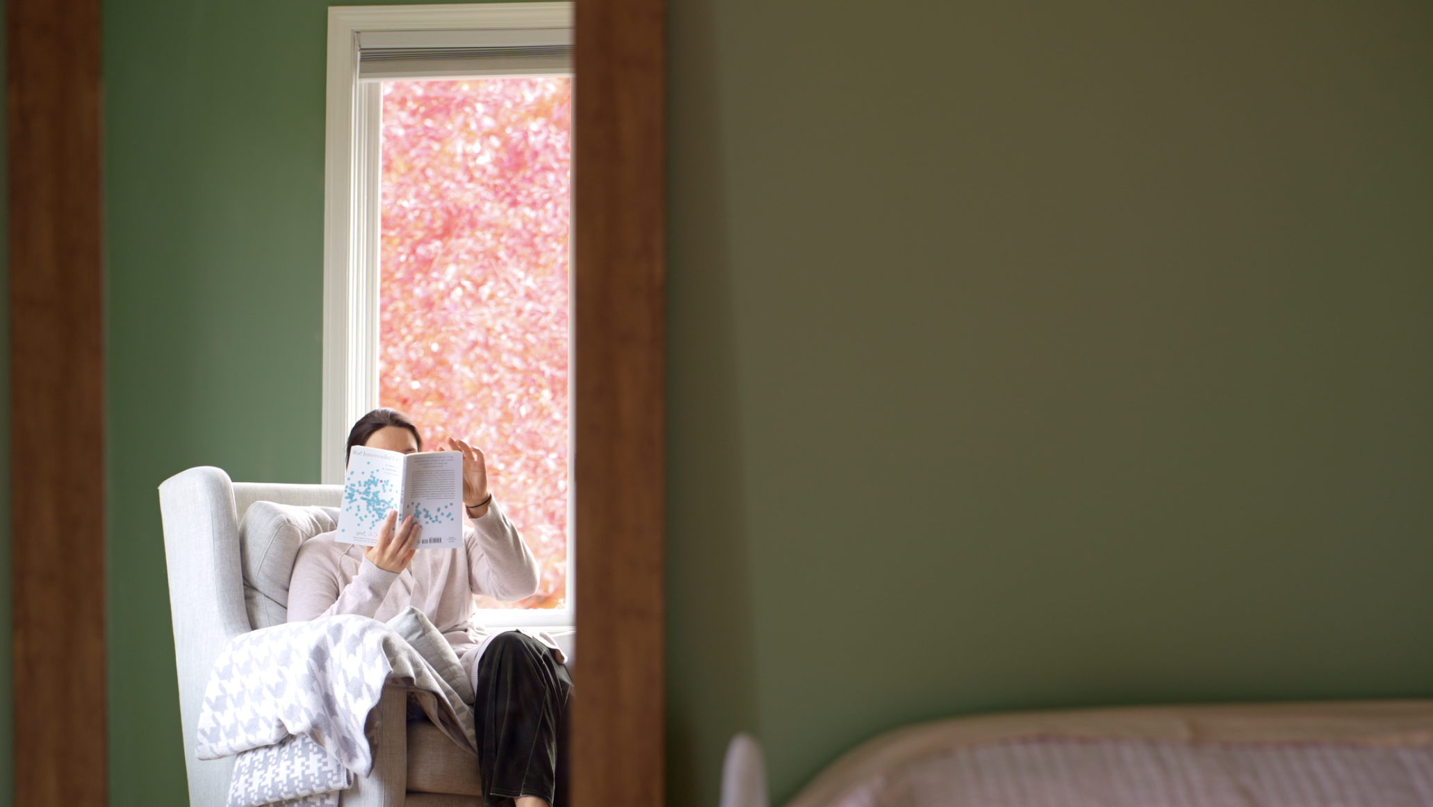 A woman sitting in a beige armchair by a window, reading a book, with autumn trees visible outside.