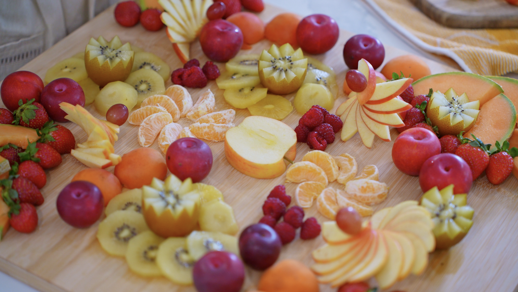 A wooden cutting board with a variety of cut and whole fruits including apples, strawberries, kiwi, oranges, raspberries, cantaloupe, and pomegranate.