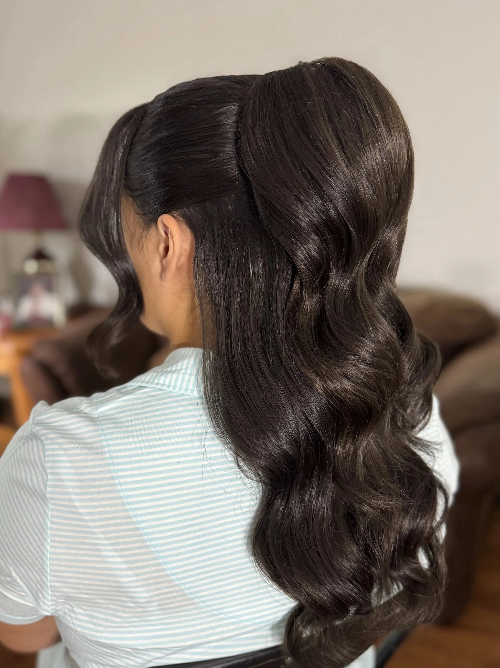 Back of a woman with long, dark, voluminous, wavy hair sitting on a chair in a room with a sofa, a lamp with a pink shade, and a photo frame in the background.