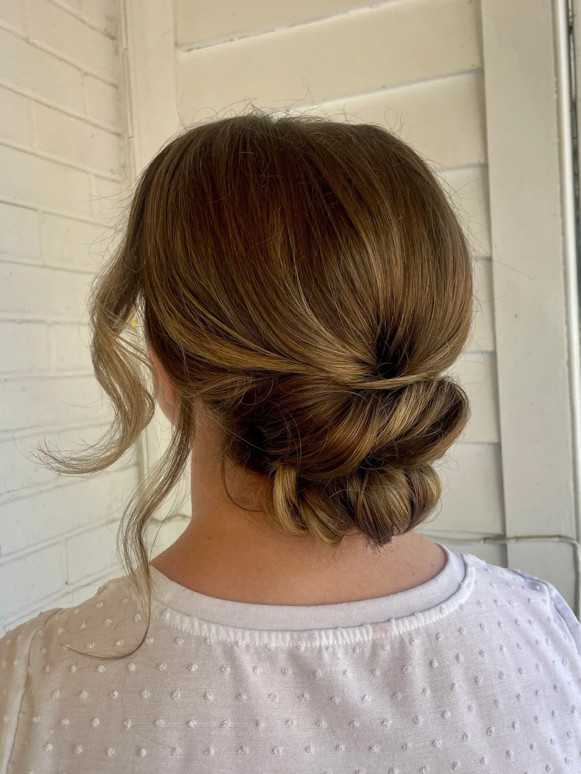 Back view of a woman with brown hair styled in an elegant updo, wearing a white textured top, standing near a white brick wall and a window.