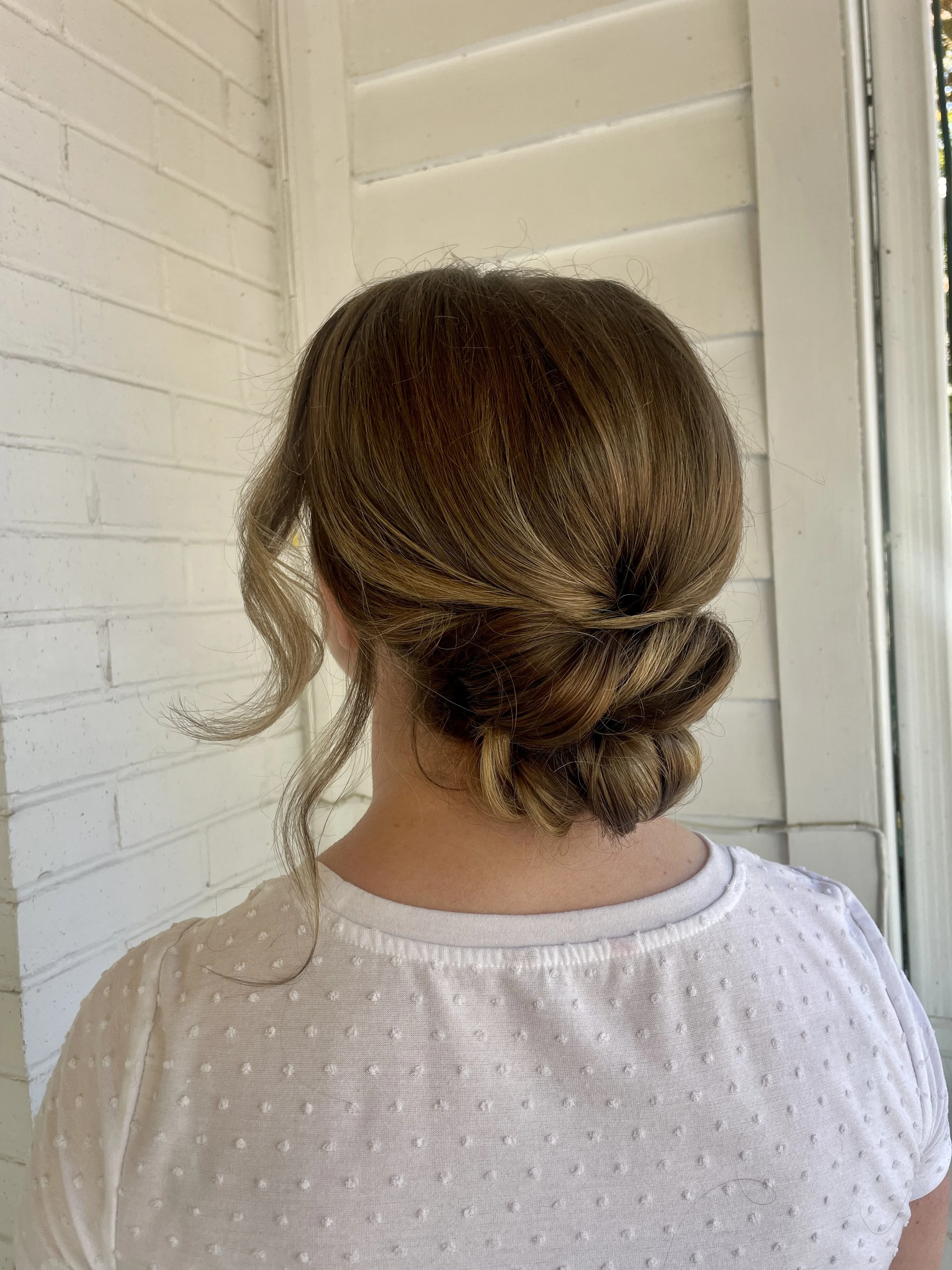 Back view of a woman with brown hair styled in an elegant updo, wearing a white textured top, standing near a white brick wall and a window.