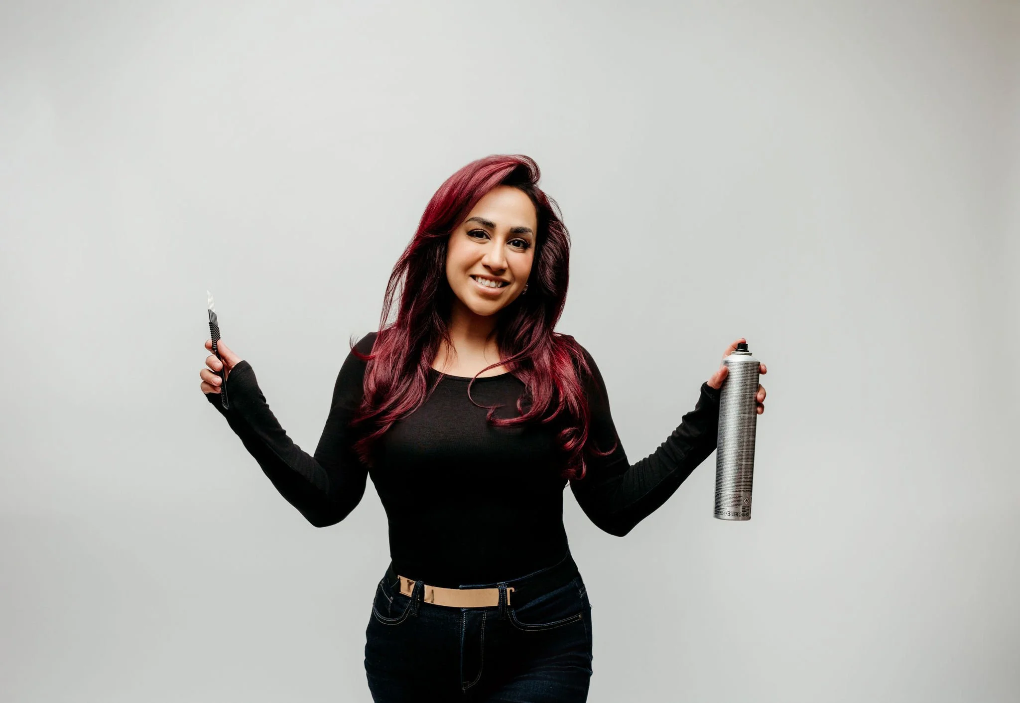 A woman with red hair and a black top, holding a hairbrush in one hand and a can of hair spray in the other, smiling at the camera against a plain light gray background.