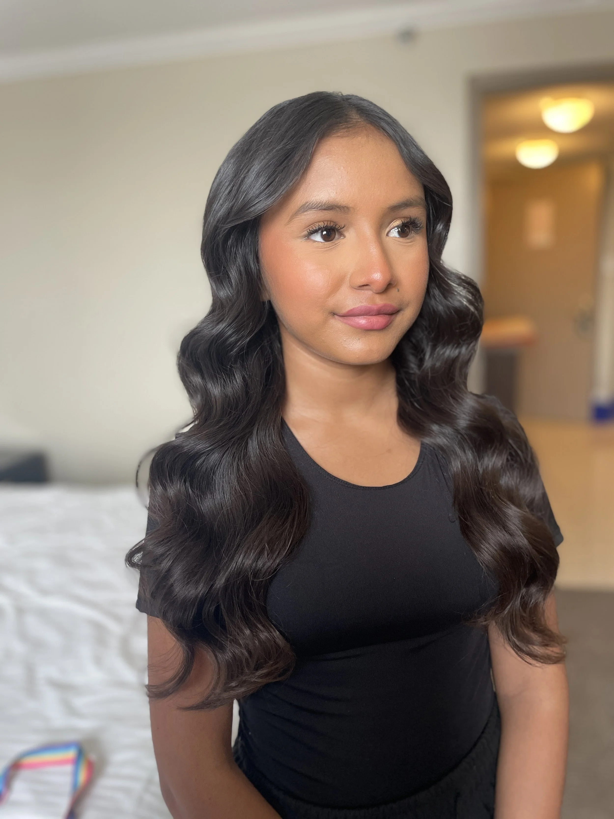 Young woman with long, wavy black hair, wearing a black shirt, standing in a room with blurred background, looking confident.