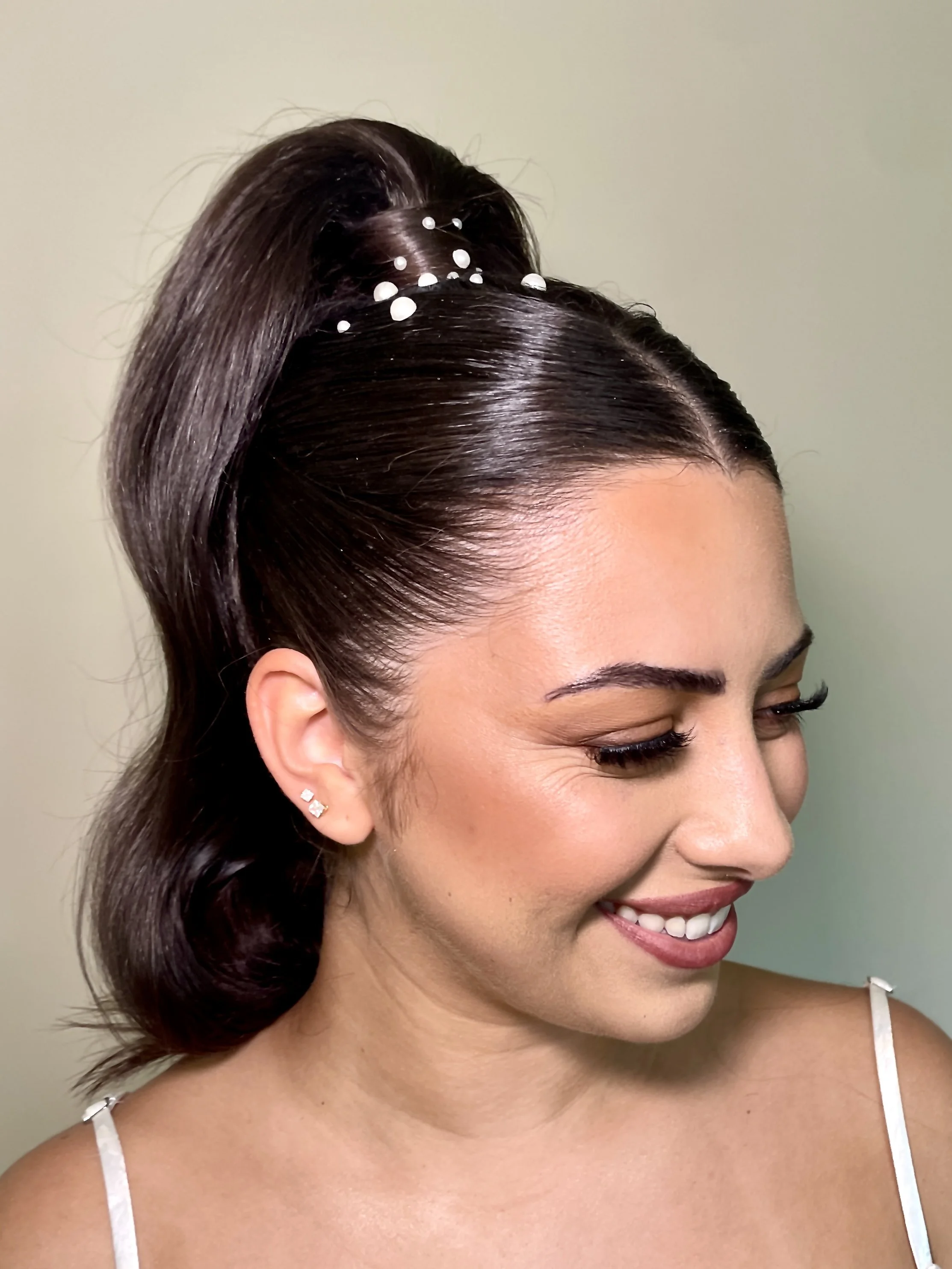 Close-up of a smiling woman with dark, shiny hair styled in an updo with white decorative beads, wearing small earrings and a white spaghetti strap top, with a neutral background.