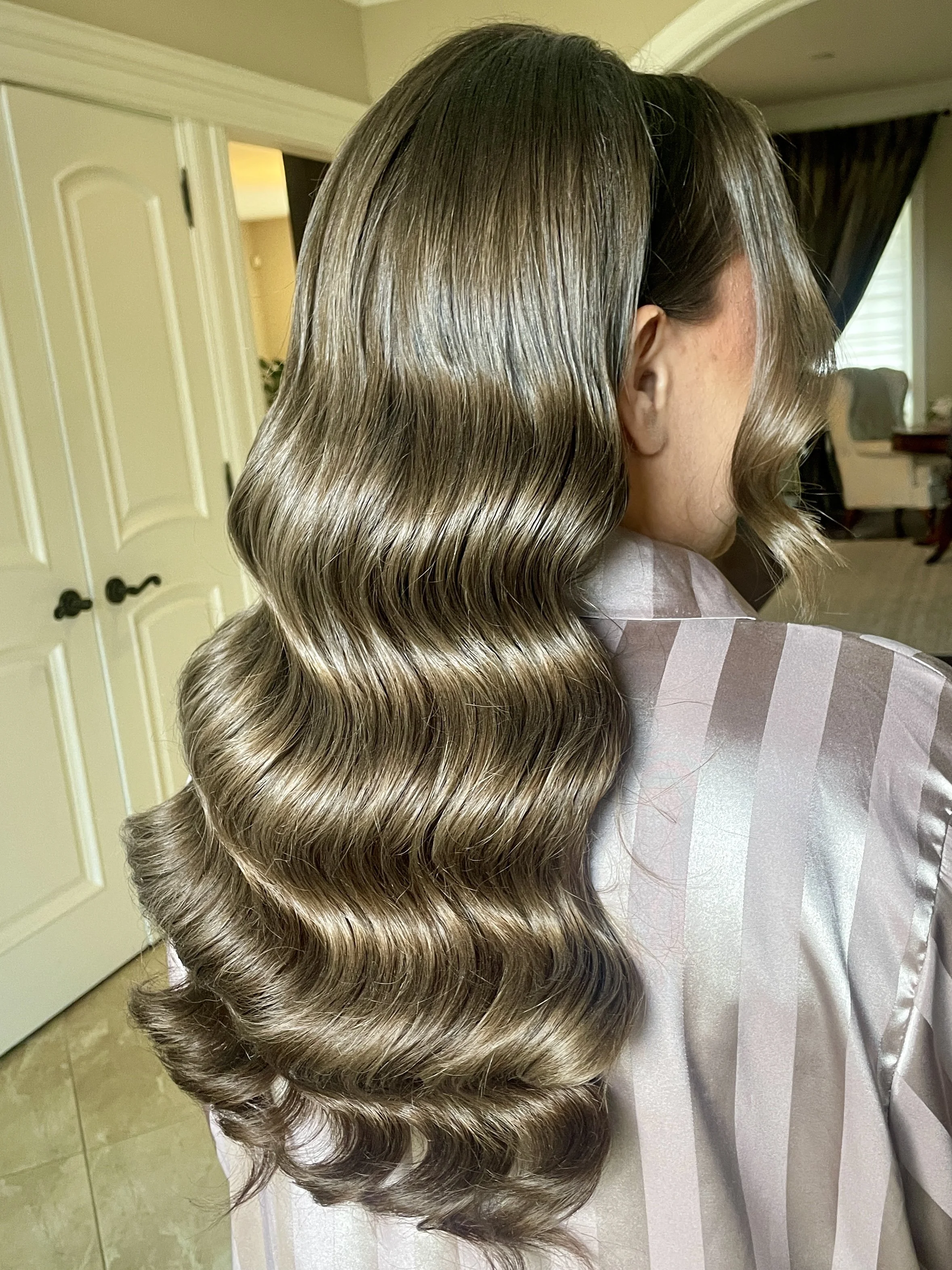 A woman with glossy, wavy, light brown hair sits in a room with a beige door and dark curtains in the background.