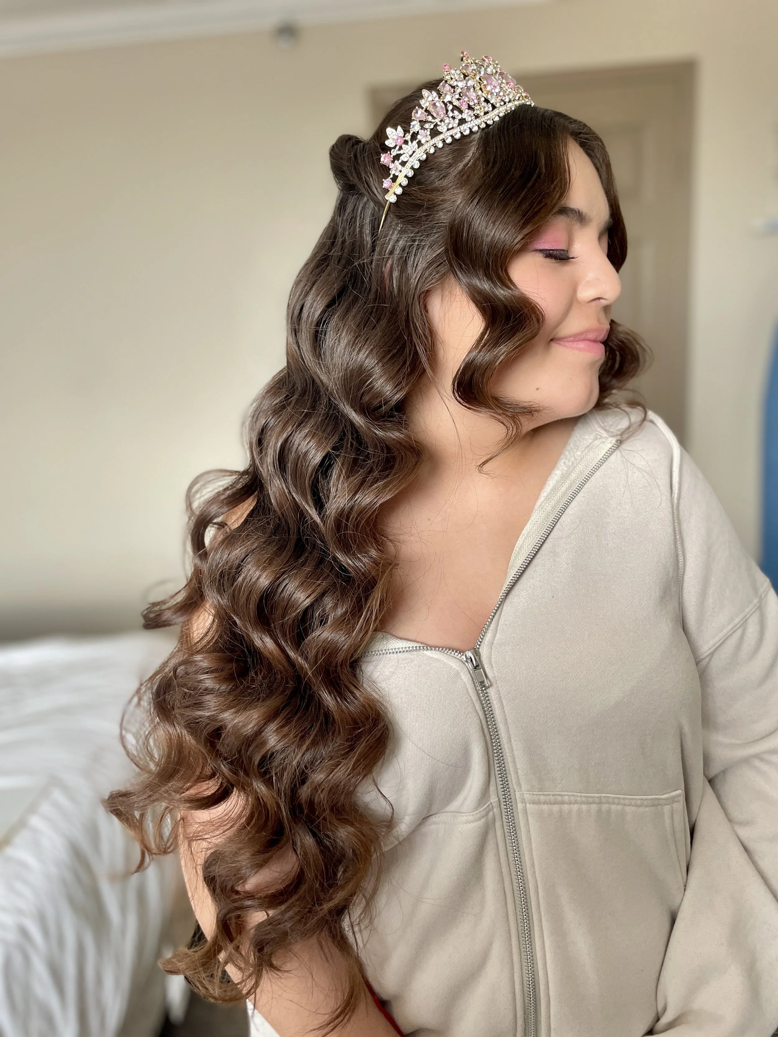 A woman with long, wavy brown hair wearing a tiara, pink eyeshadow, and a beige zip-up hoodie.