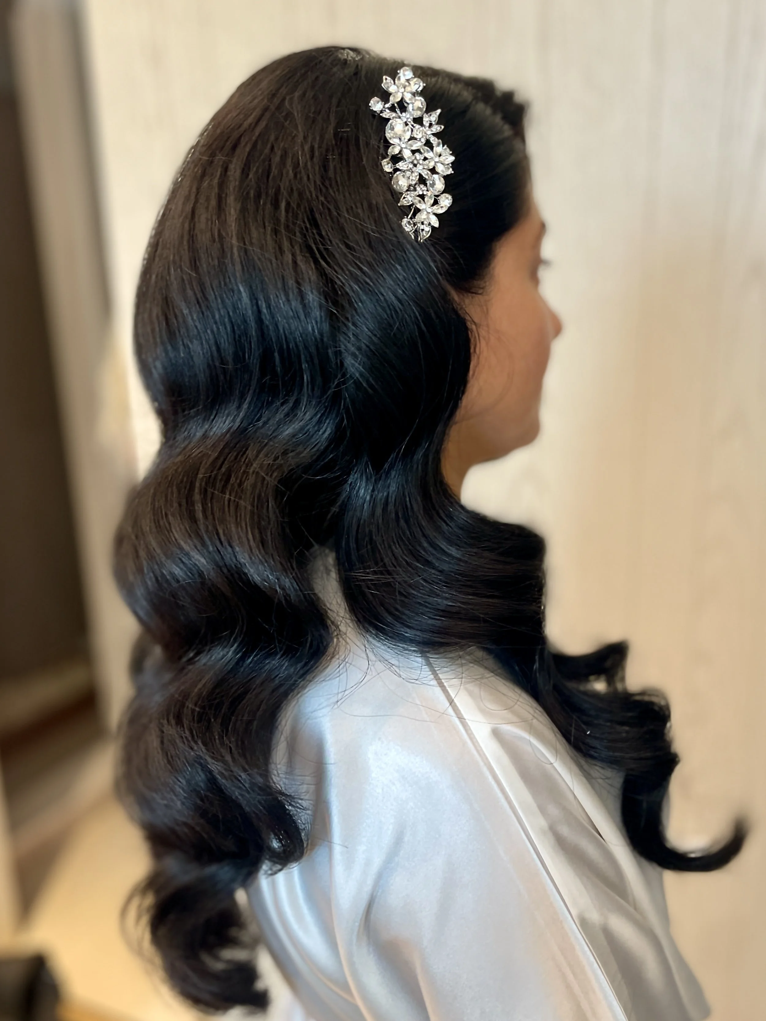 A woman with long, shiny black hair styled in loose waves, wearing a silver hair accessory with floral design.
