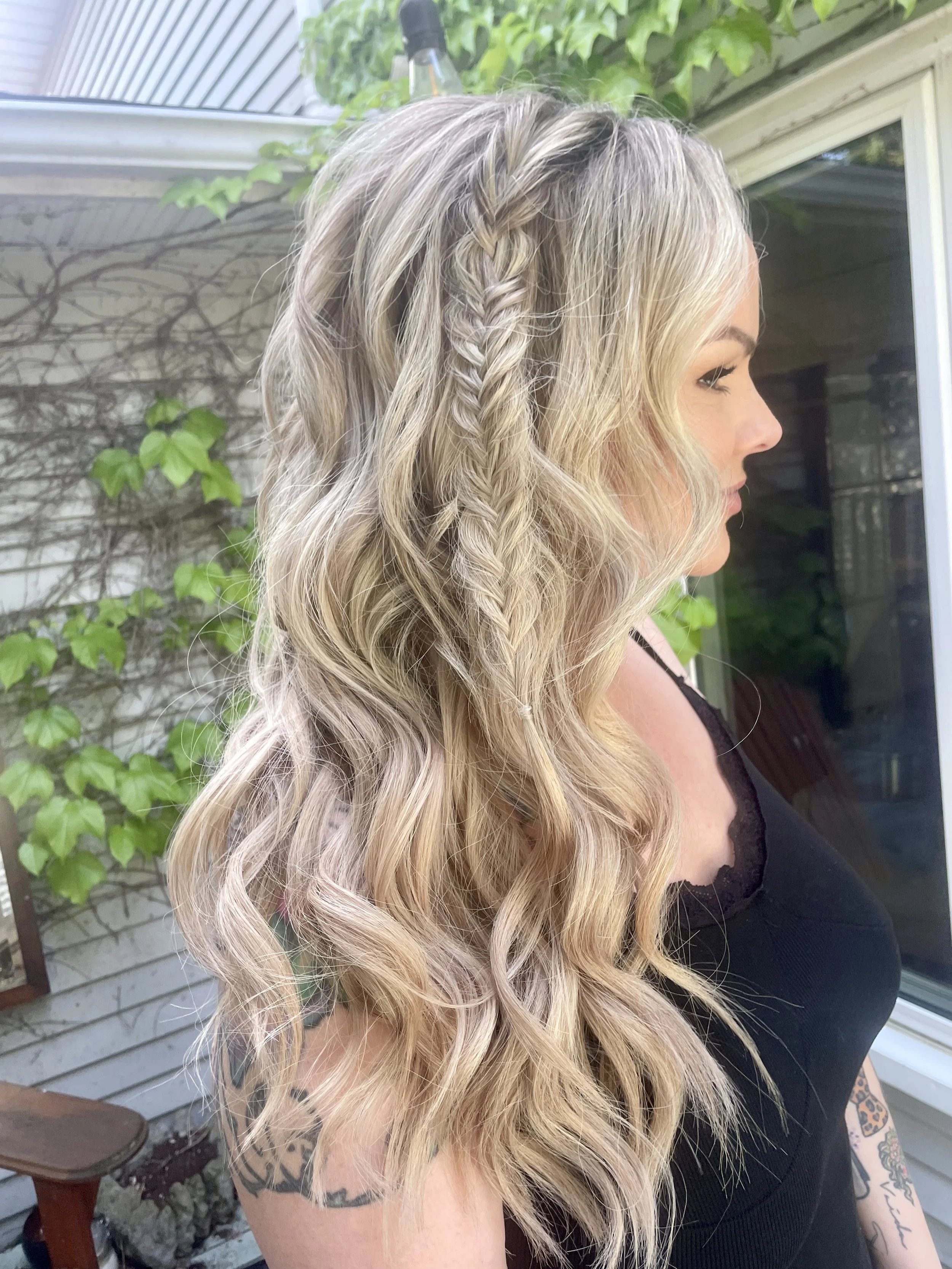 A woman with long, wavy blonde hair, featuring a small braid on the side, standing outdoors near a house with a sliding glass door, green leaves, and a vine growing on the wall.
