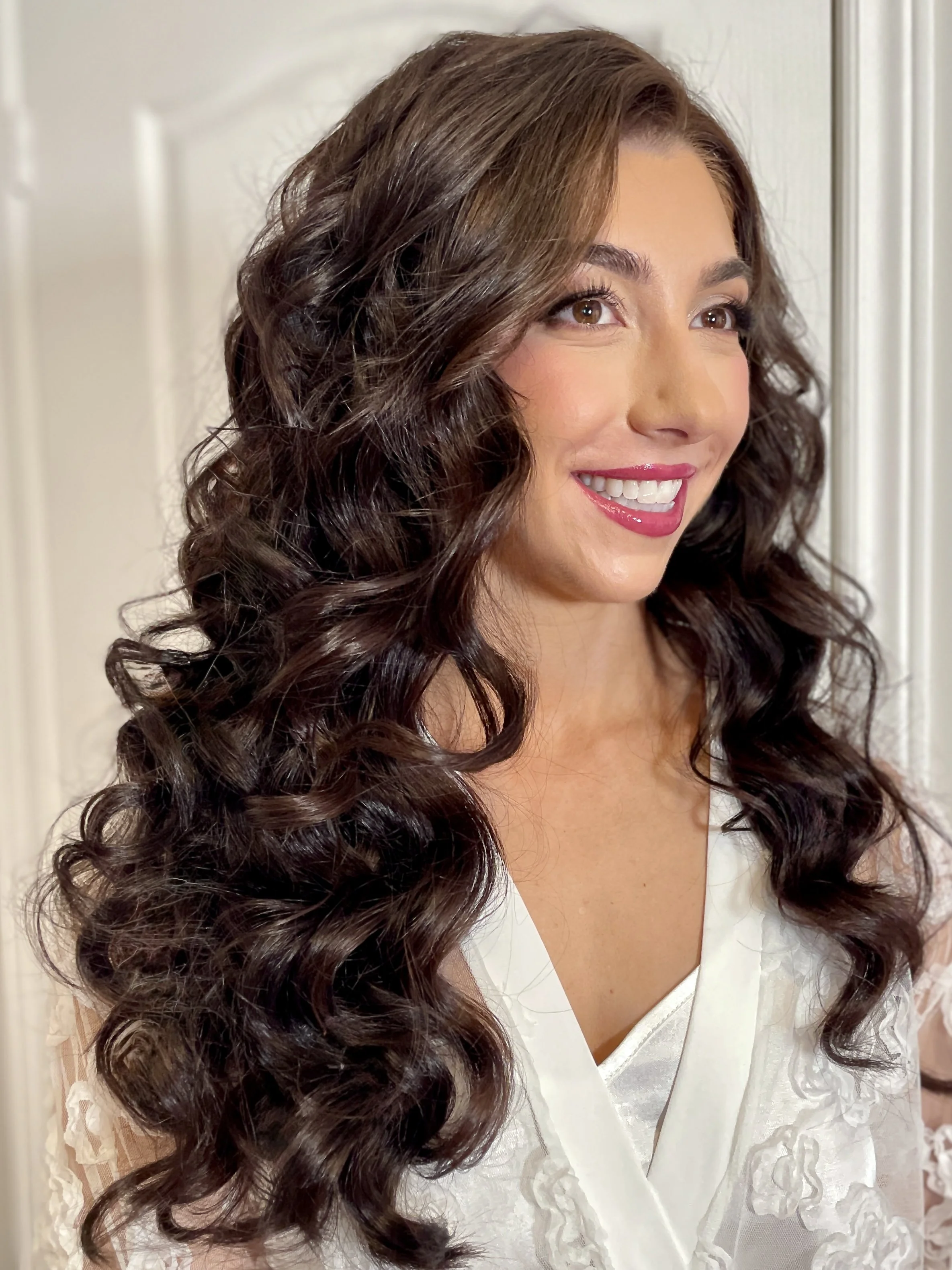 A woman with long, dark, curly hair smiling and wearing a white top.