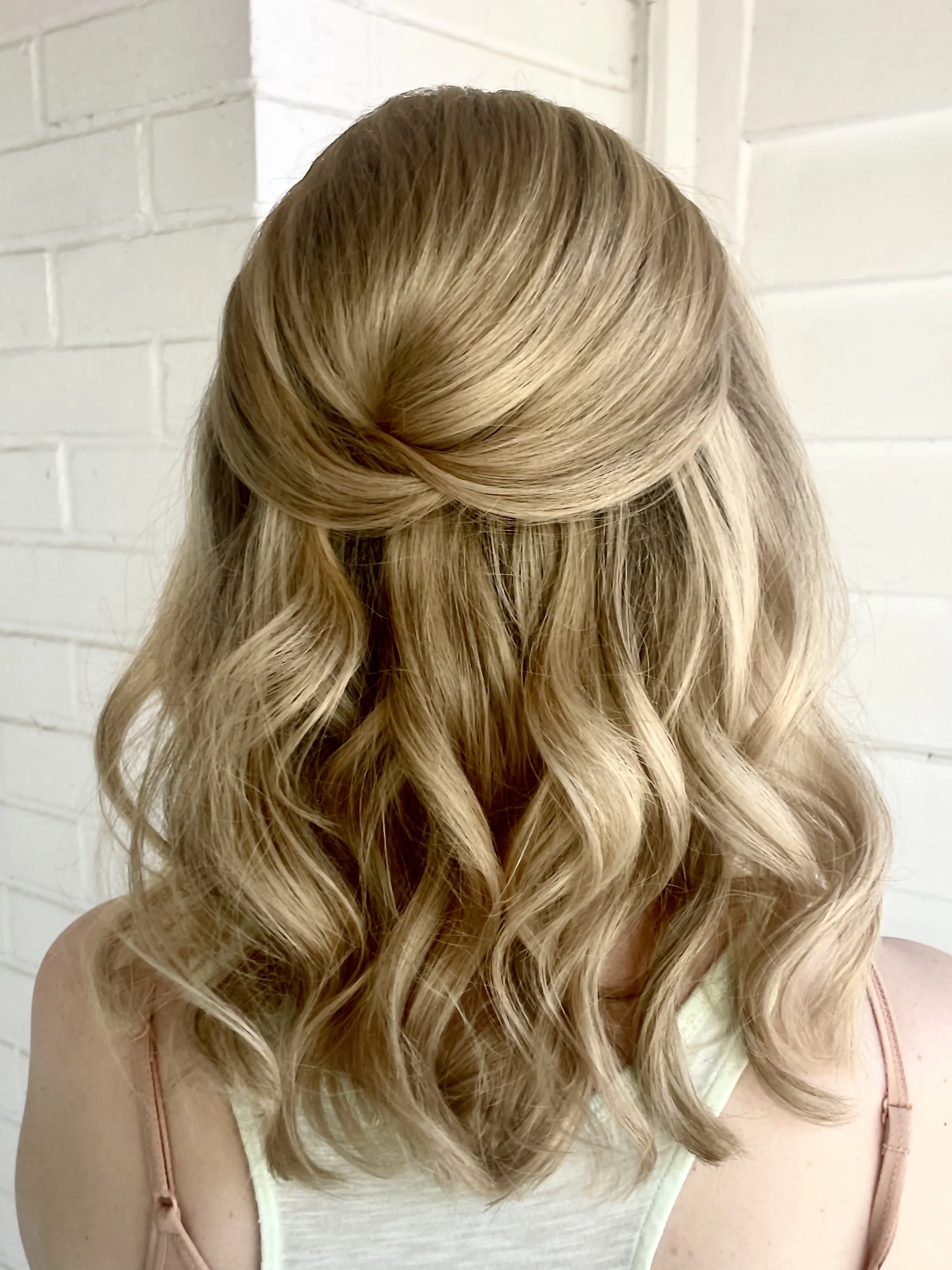 Back view of a woman's hair styled in loose waves with a twisted section at the crown, set against a white brick wall.