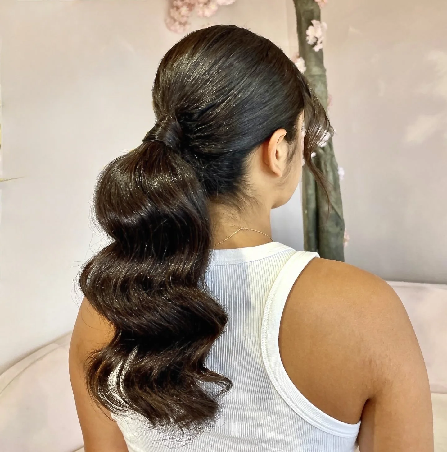 Back view of a woman with wavy dark brown hair styled in a loose low ponytail, wearing a white sleeveless top and a delicate gold necklace, standing in front of a light-colored wall with a floral decoration.