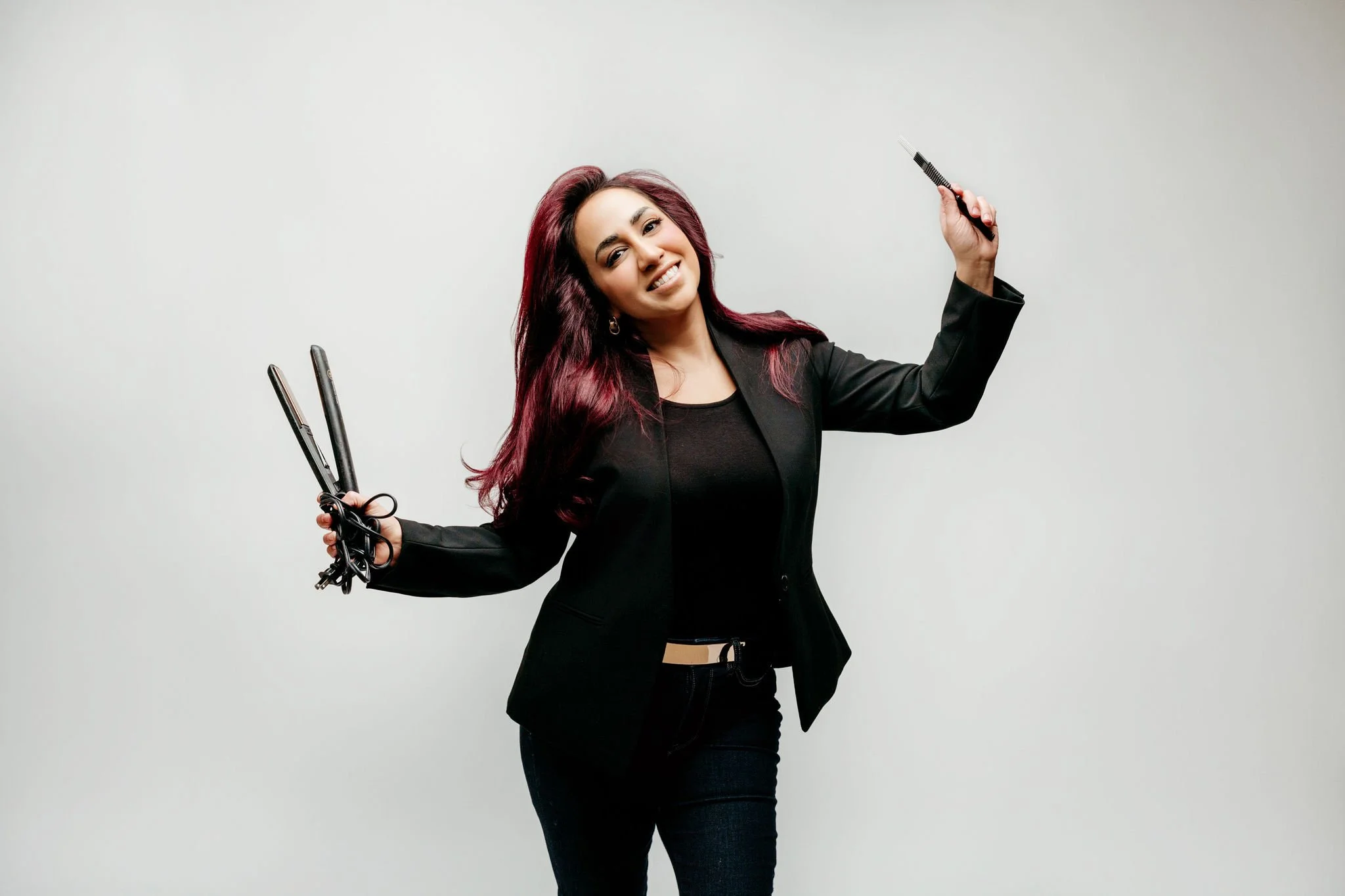 Woman with burgundy hair smiling and holding a curling iron in one hand and a flat iron in the other, standing against a plain white background.