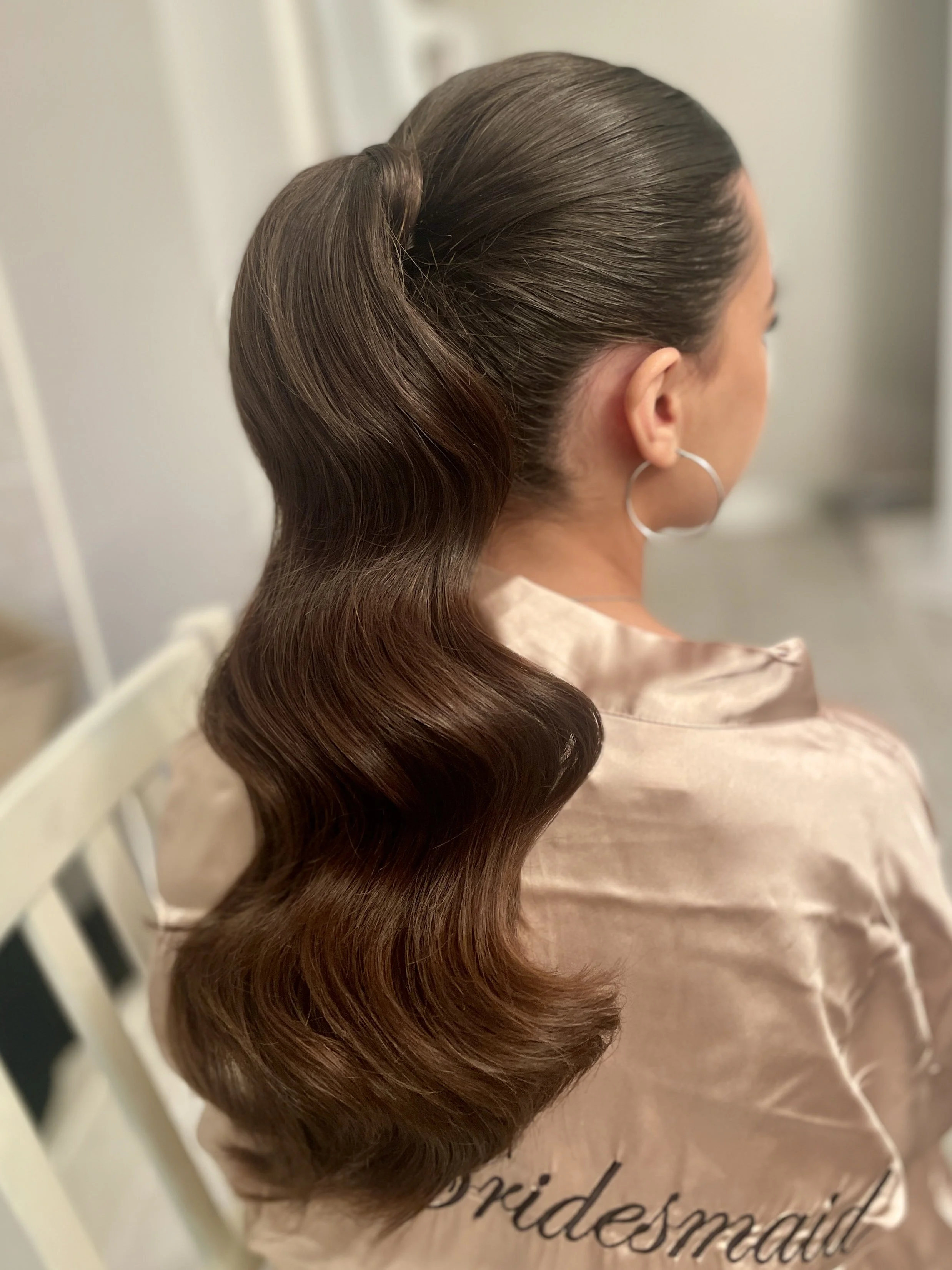 A woman with dark brown, wavy hair styled in a high side ponytail, wearing earrings and a satin robe.