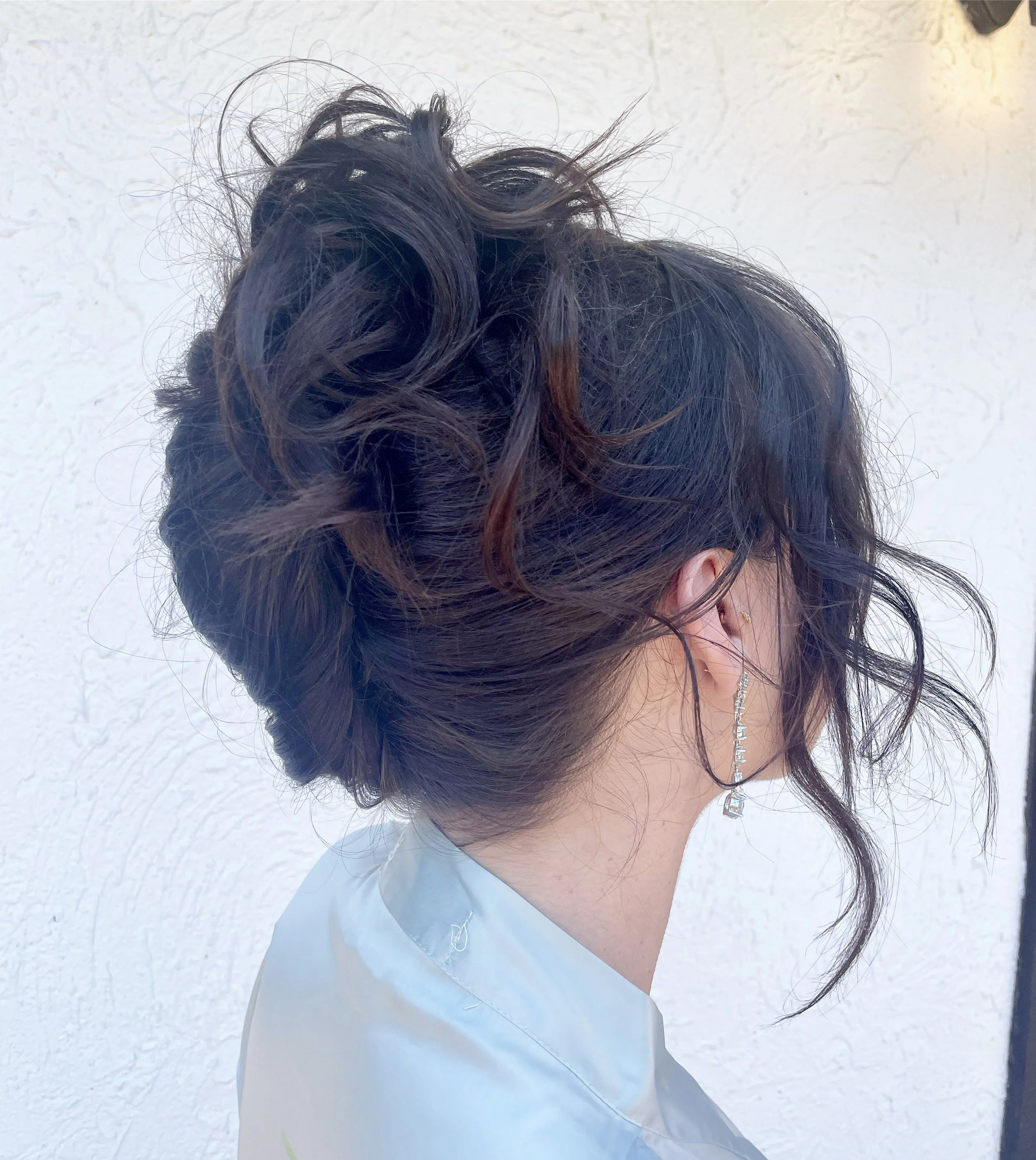 Side view of a woman with her hair styled in an elegant updo with loose curls and wisps, wearing a white top and an earring, standing against a textured white wall.