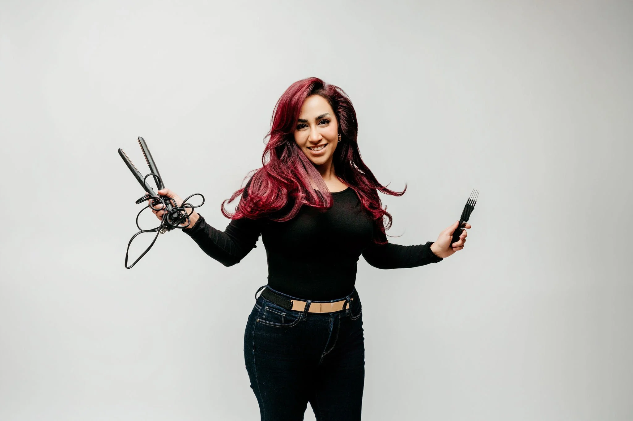 Young woman with red hair holding a flat iron and hairbrush in a studio with a plain white background.