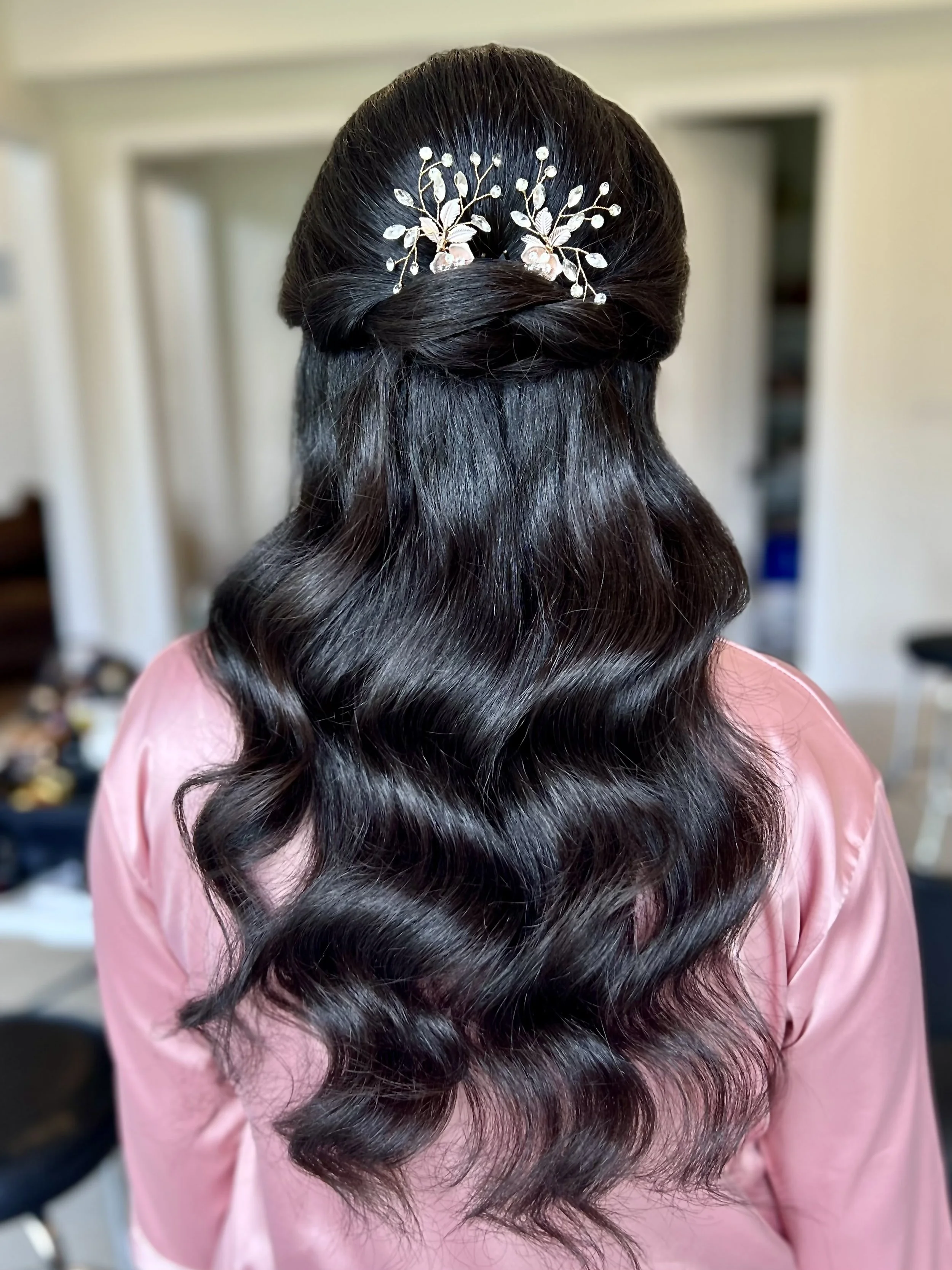 A woman with long, dark, wavy hair styled with loose curls, wearing a pink satin robe, and a decorative hairpiece with pearls and flowers