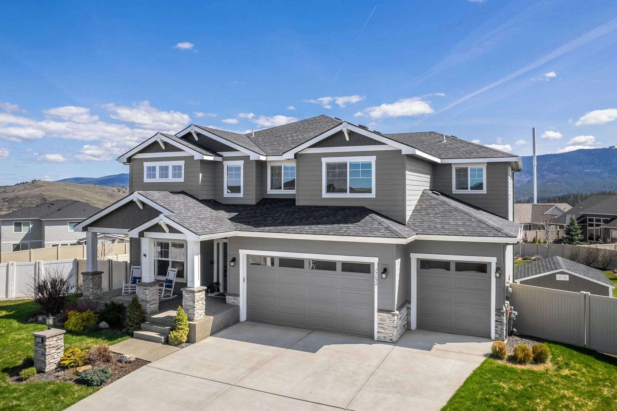 A modern two-story house with gray siding, a multi-gabled roof, and a three-car garage, surrounded by a green lawn and landscaping, with mountains in the background.