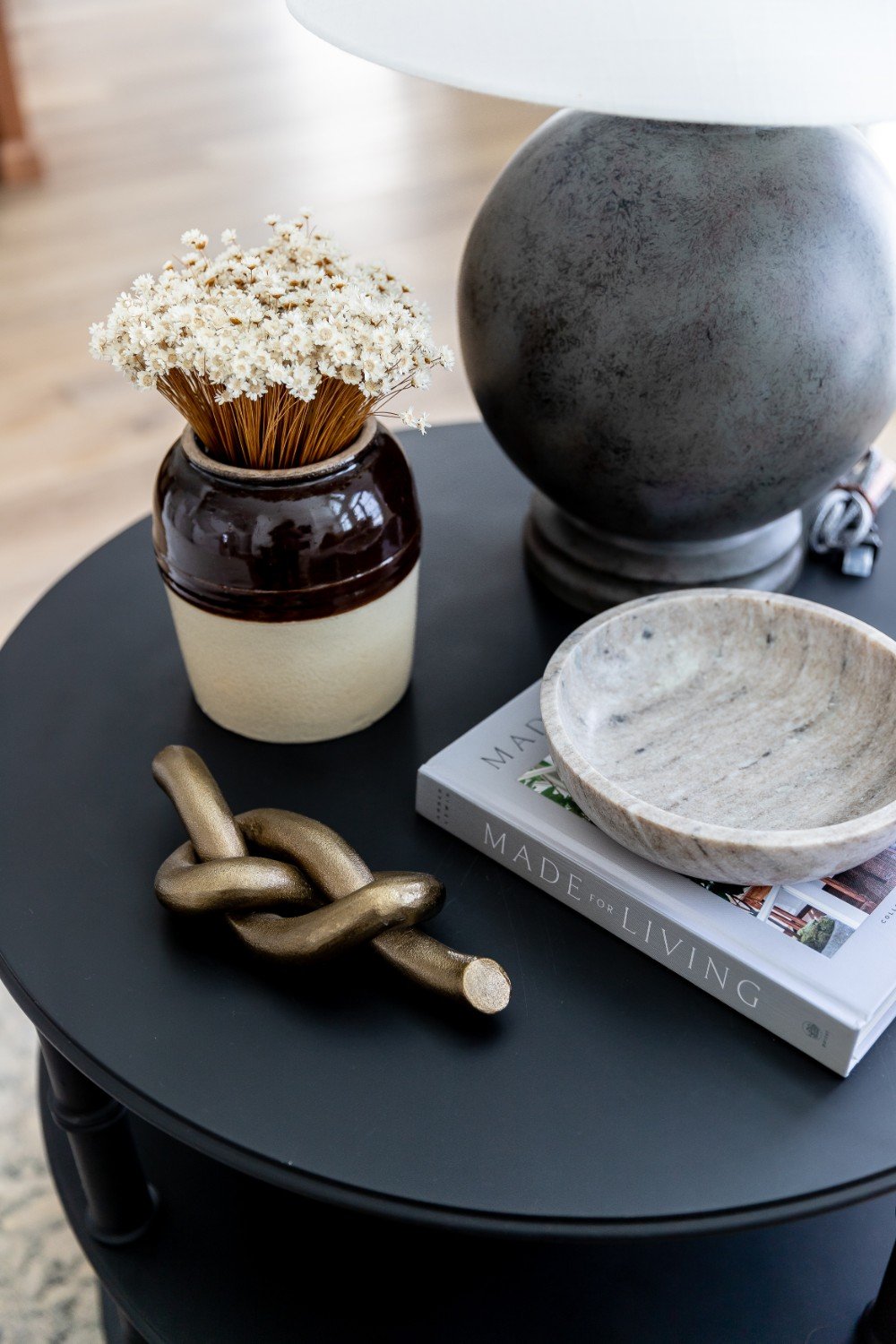 Decorative table with dried flowers in a ceramic vase, a stone bowl, a book titled "Made for Living," a brass knot sculpture, and a large stone lamp, all arranged on a black round table.