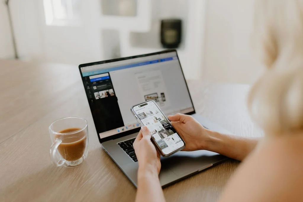 Person holding a smartphone in front of a laptop while sitting at a wooden table with a cup of coffee, in a bright room.