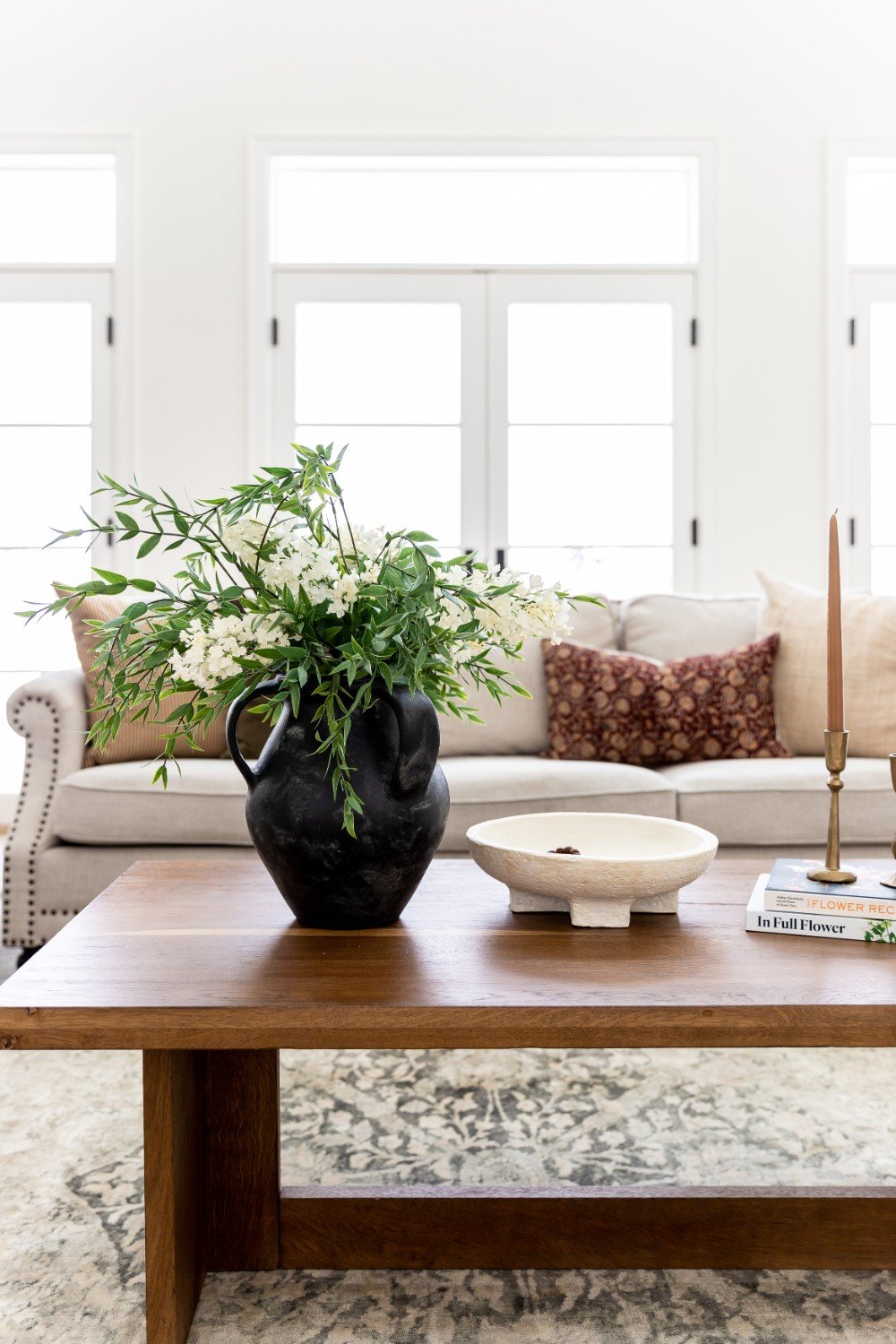 Living room with a wooden coffee table featuring a black vase with white flowers and green foliage, a white ceramic bowl, a candle holder with a pink taper candle, and books, with a beige sofa adorned with patterned and plain throw pillows behind.